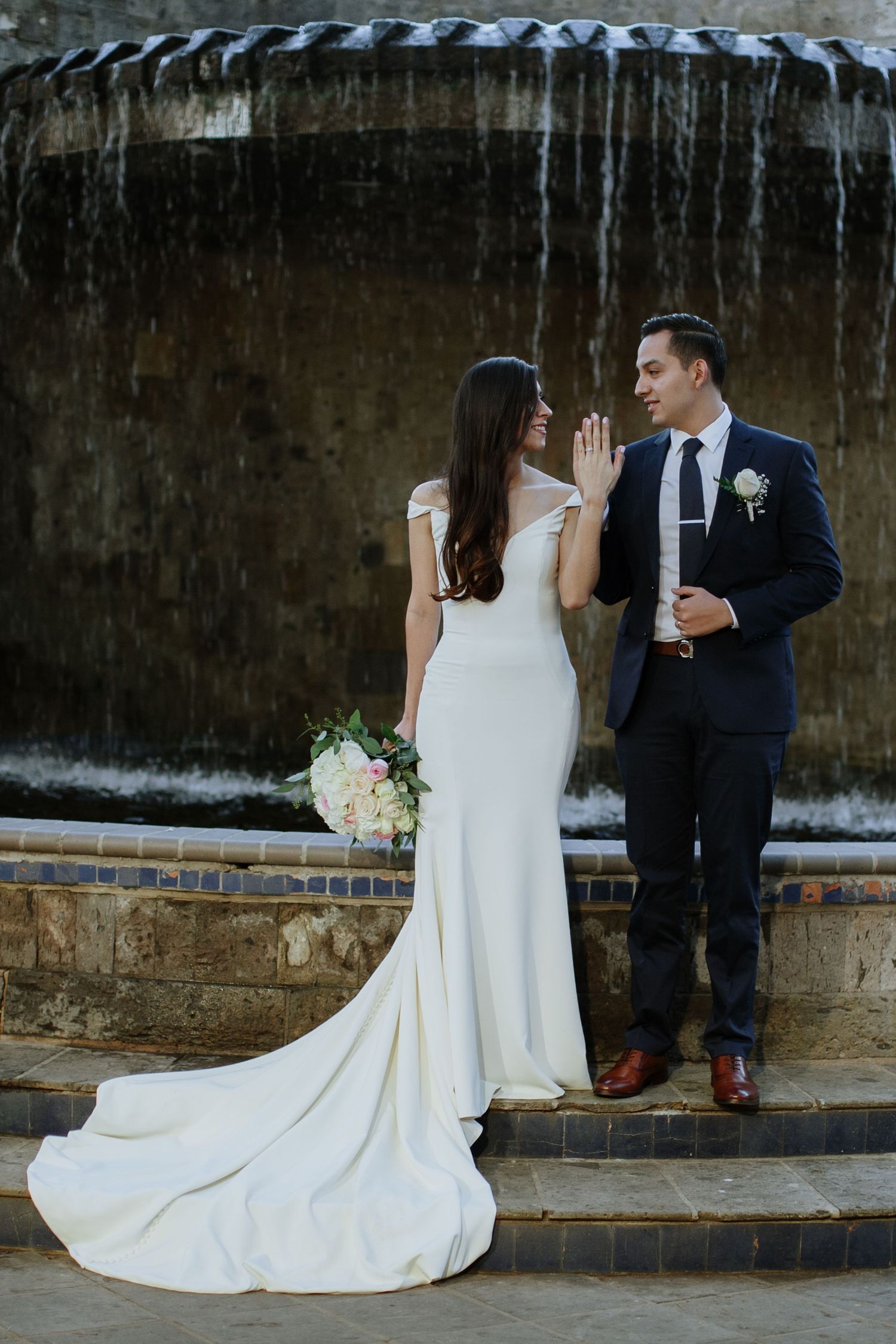 Bride and groom pose together in front of a stone fountain, bride holding a blush bouquet in elegant white gown.
