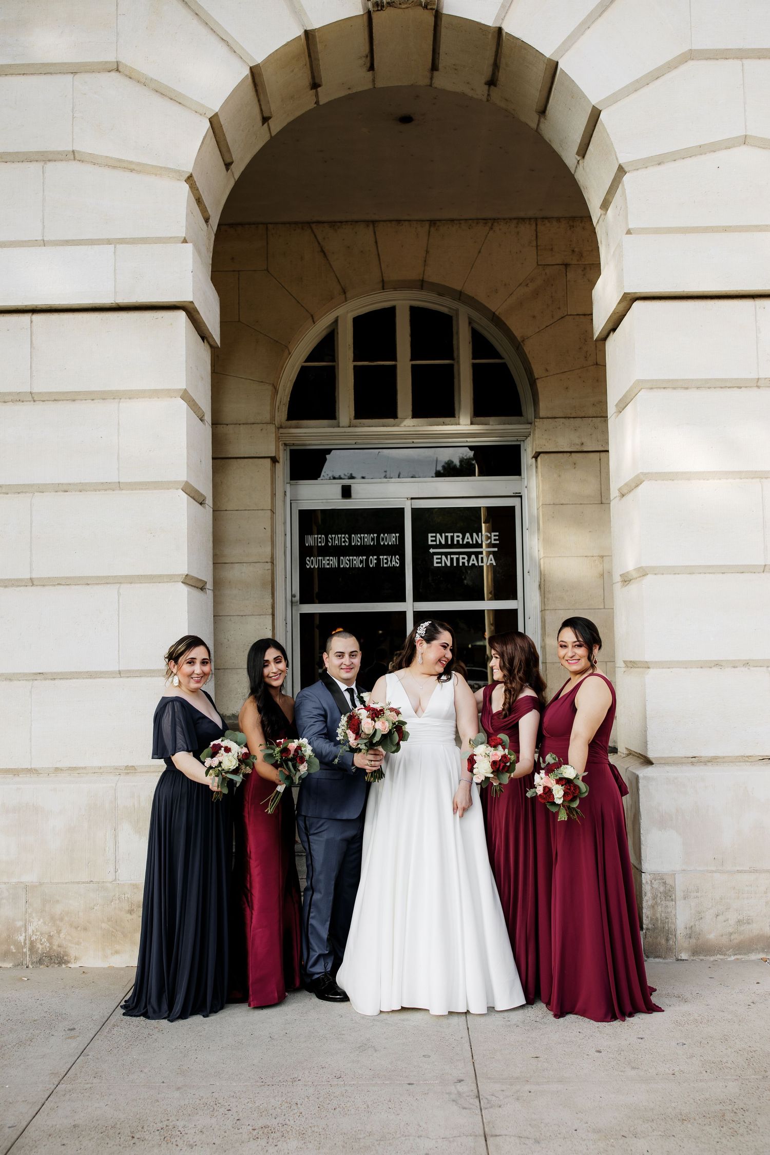 Bride and groom pose with bridesmaids in burgundy and navy gowns outside a grand stone building entrance.