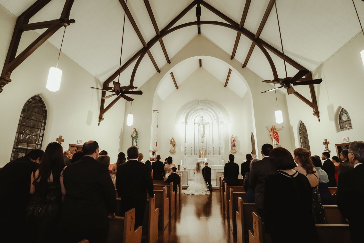 Guests stand during traditional wedding ceremony inside beautiful white Gothic church with wooden pews.