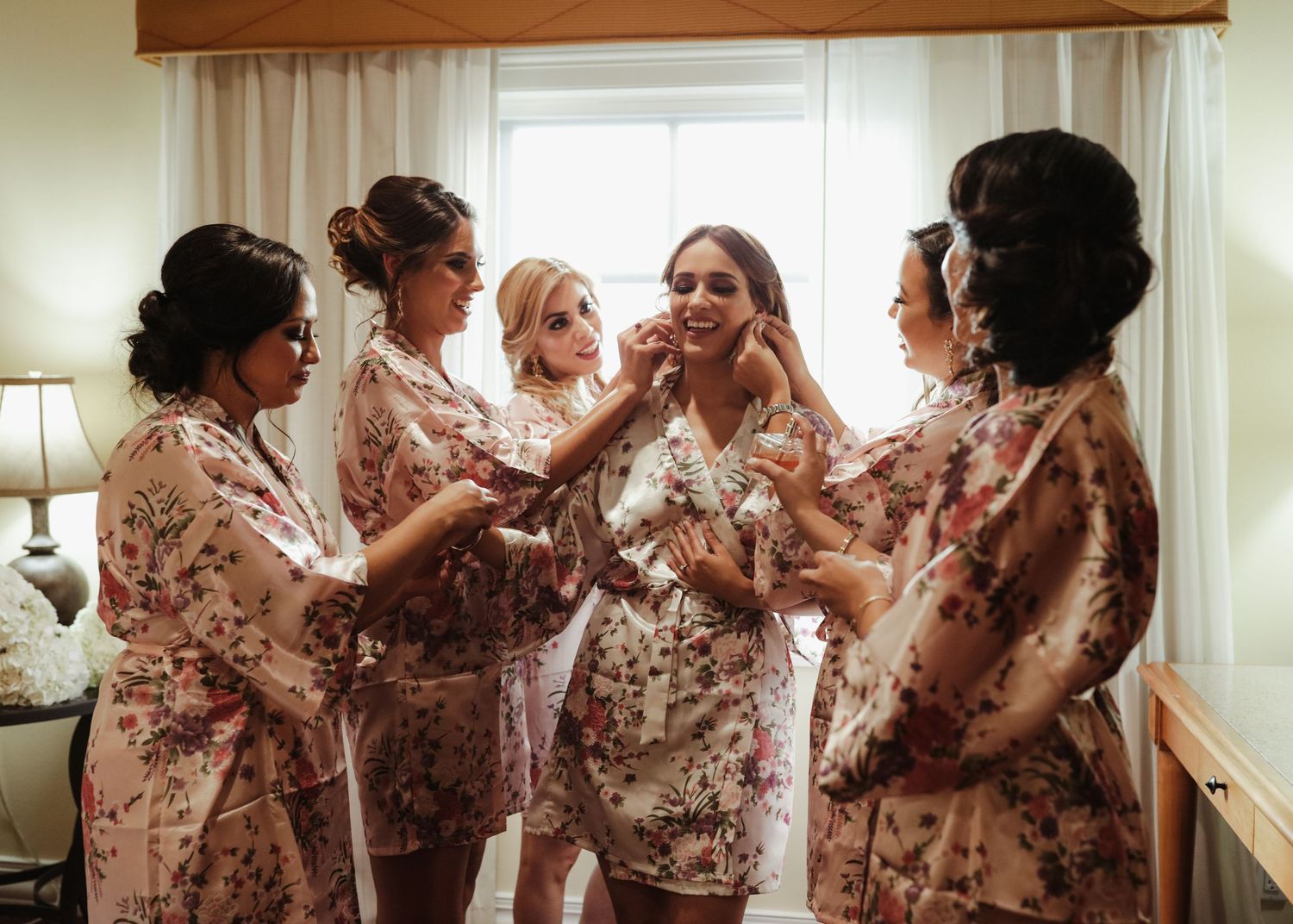 Bridesmaids in floral robes helping bride with earrings during wedding morning preparations.