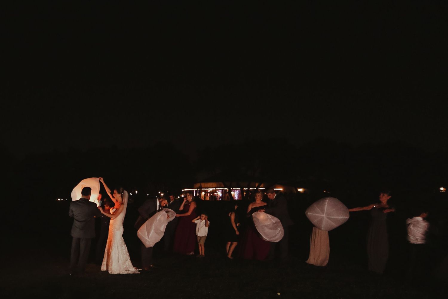 Bride, groom, and wedding guests hold white paper lanterns under dark night sky.