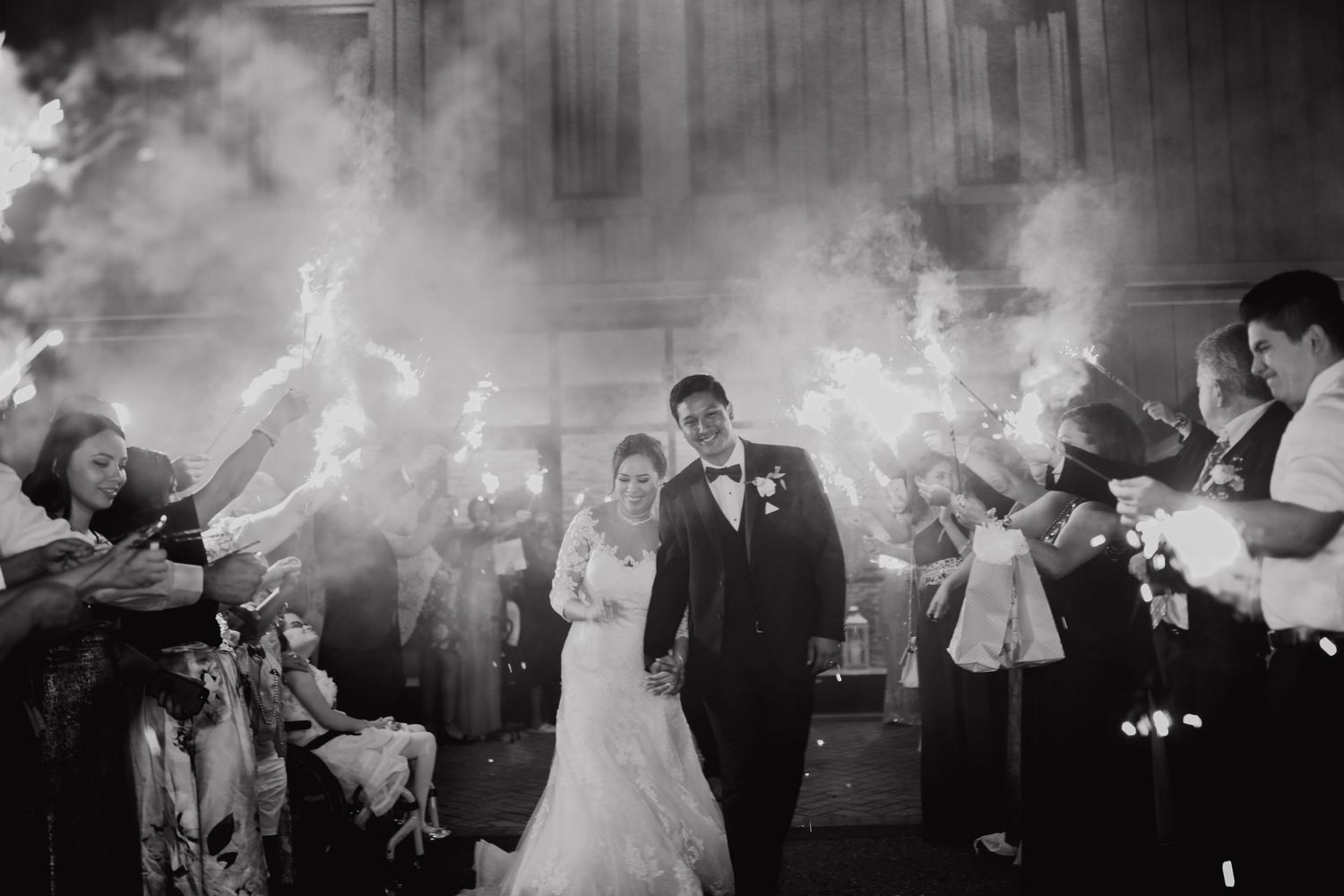 Black and white photo of bride and groom exiting through sparkler tunnel with guests cheering.