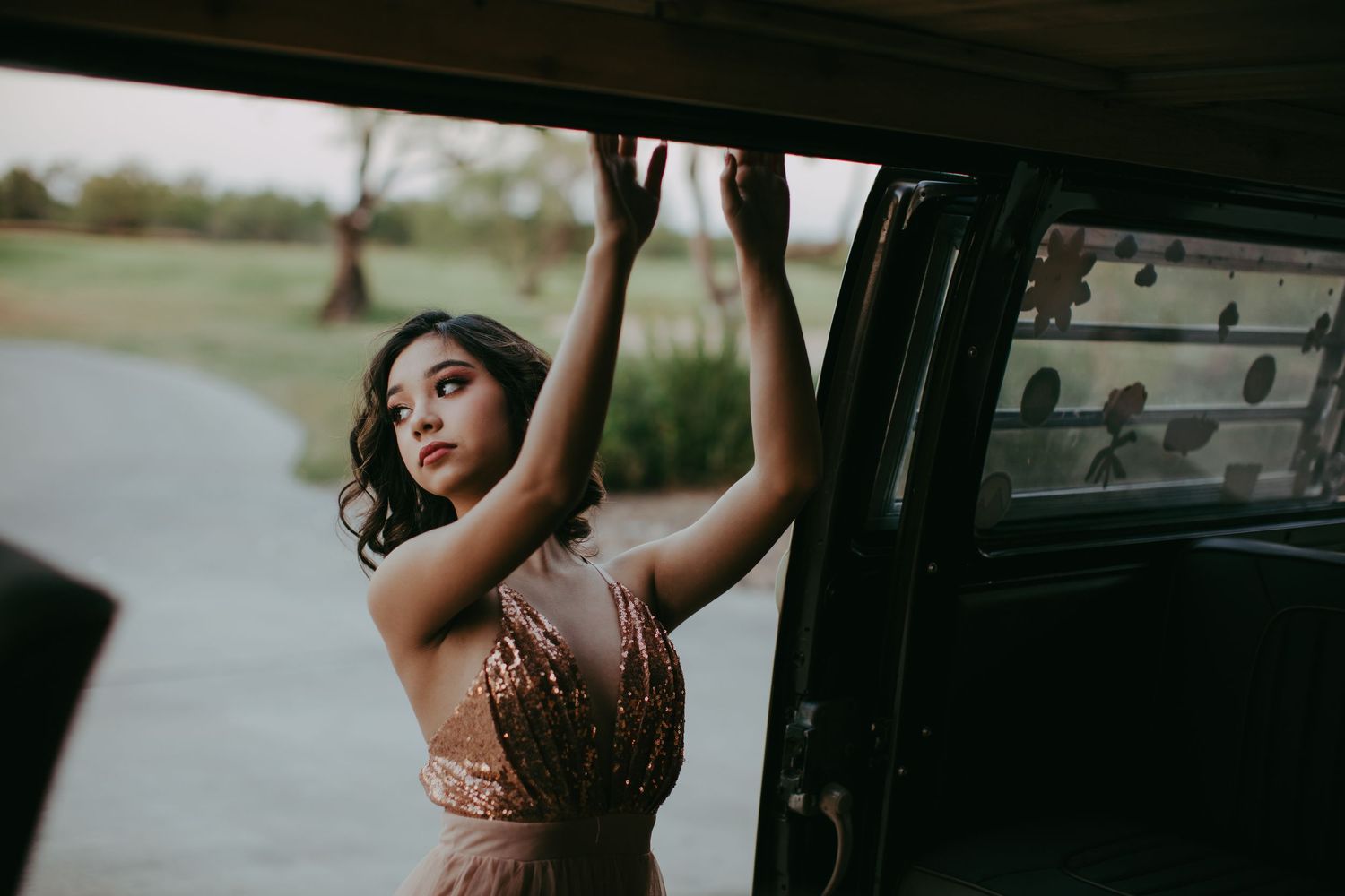 A person in a formal evening dress poses next to an open car door against a moody outdoor backdrop.