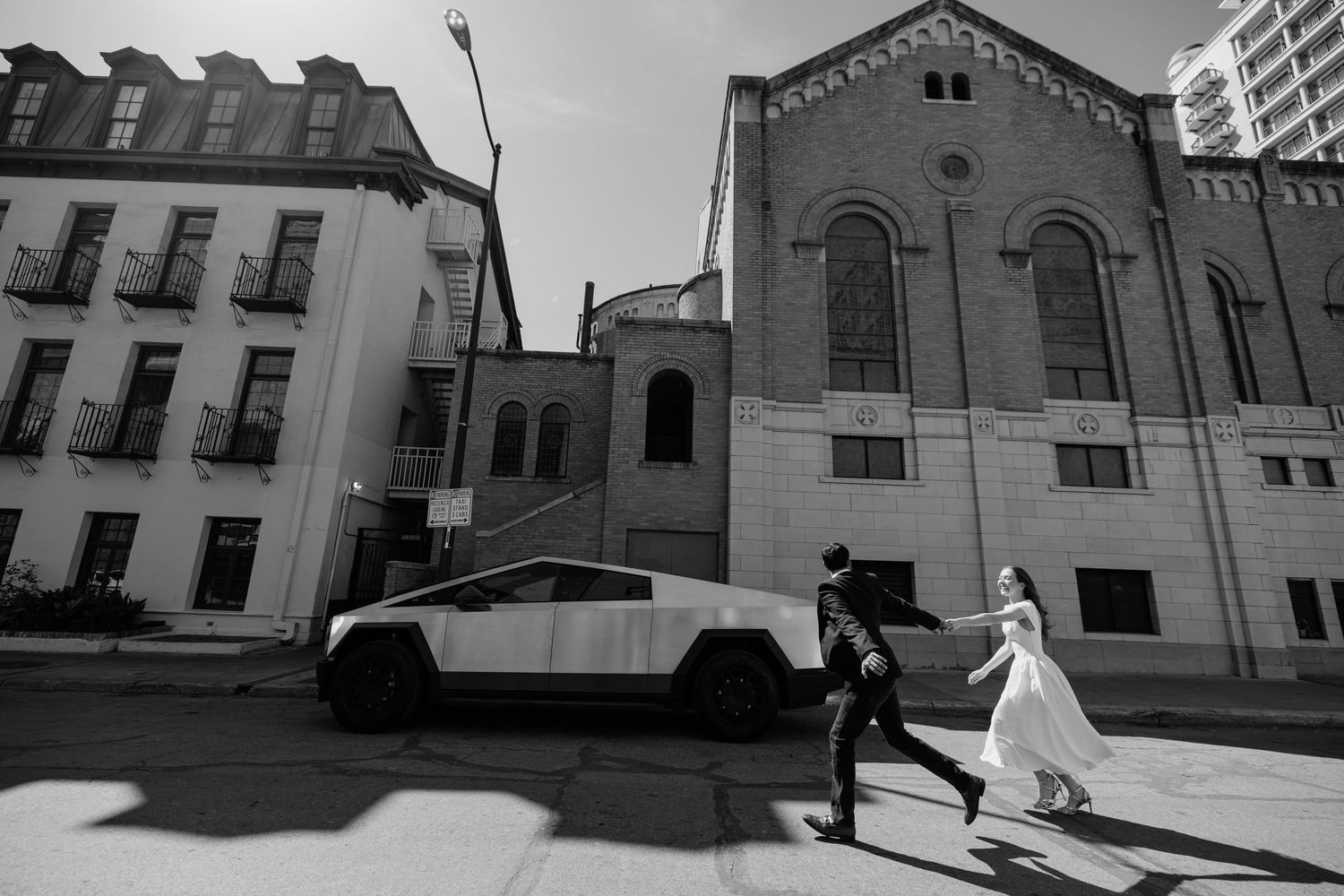 Black and white wedding photo of bride and groom running past a Tesla Cybertruck on a city street.