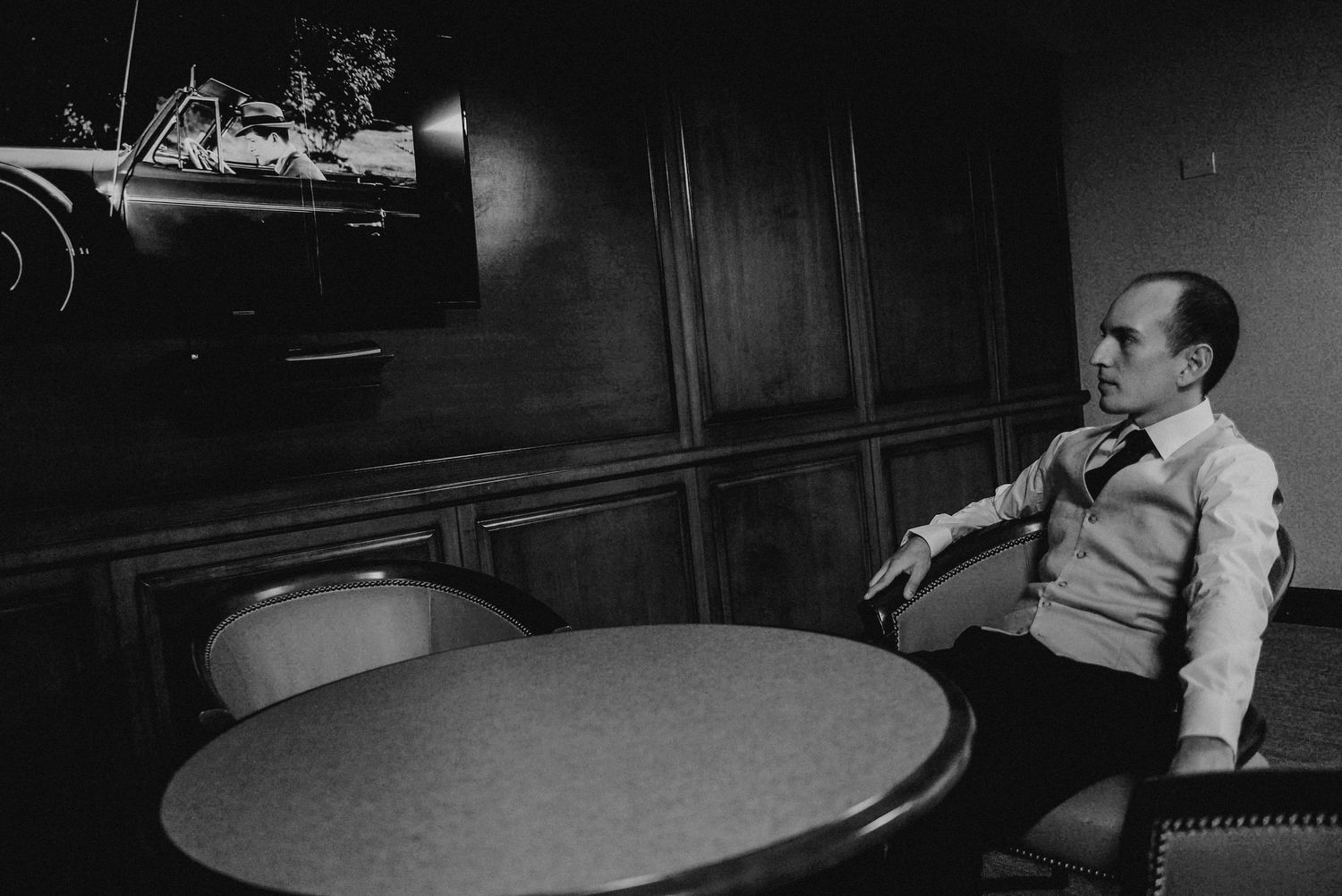 Black and white photo of man sitting alone at round table in dimly lit room.