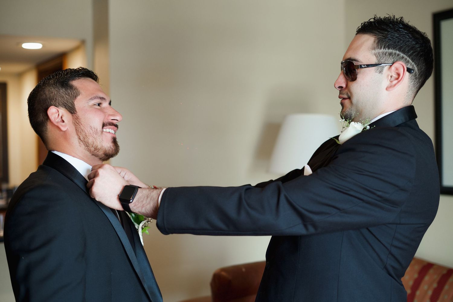 Groomsman adjusts boutonniere on smiling groom in black suit before wedding ceremony.