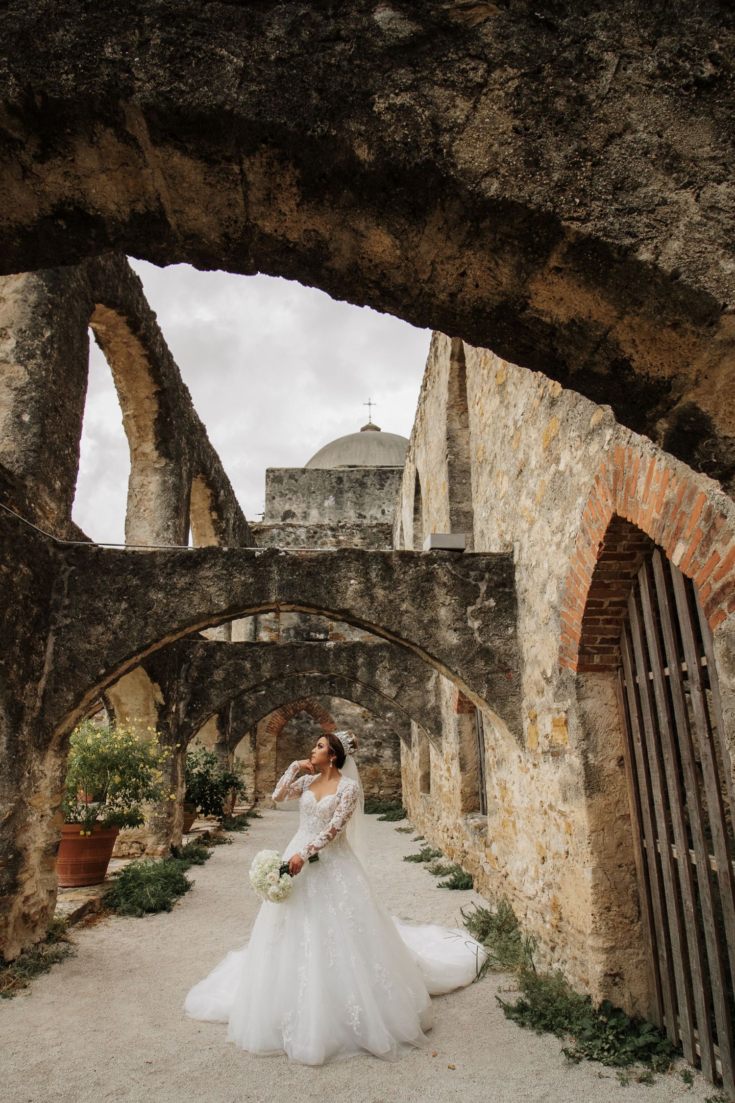 Bride in a flowing lace gown posing among stone arches at a historic mission on her wedding day.