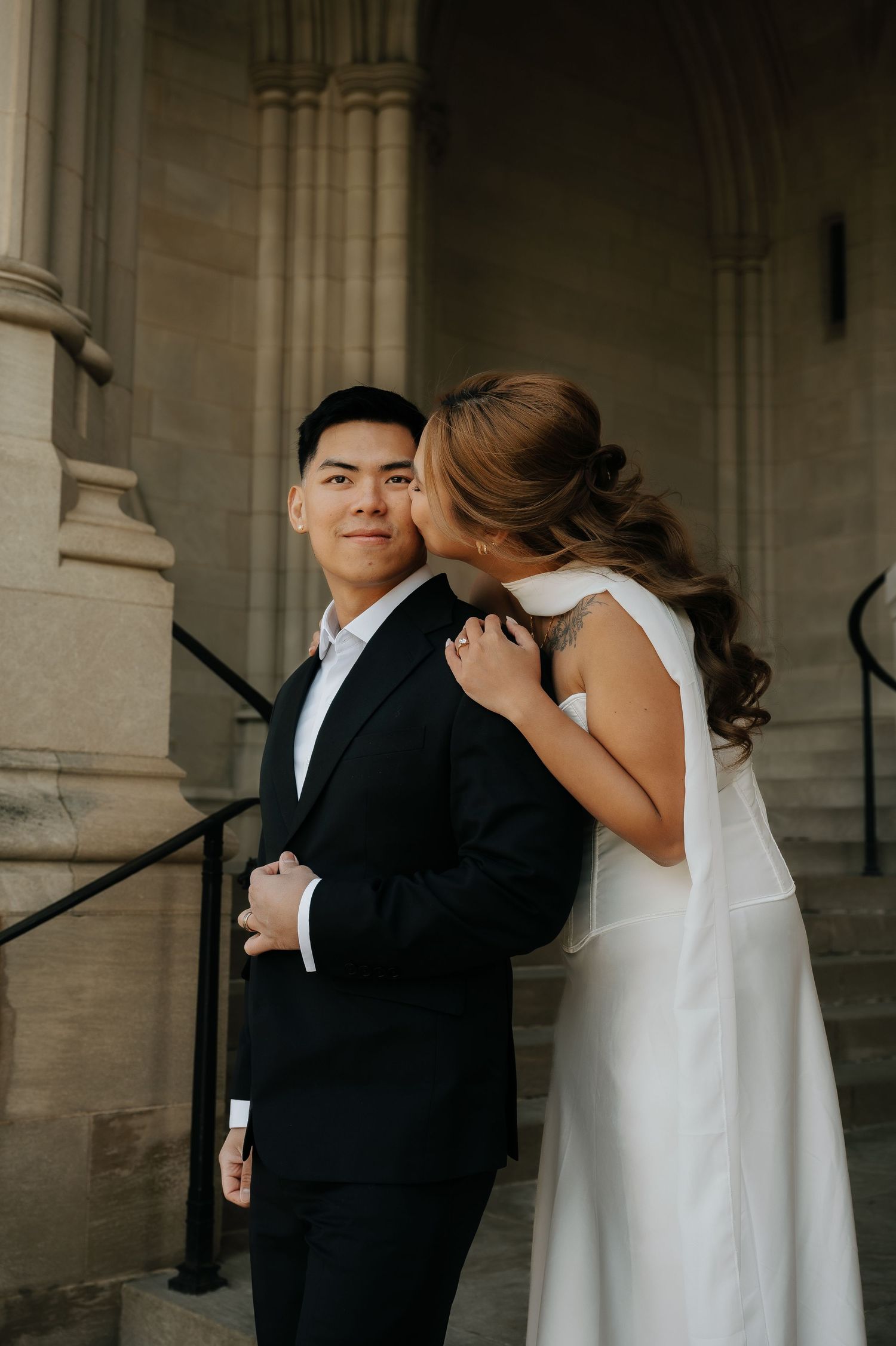 Bride in white dress kisses groom in black suit on cheek outside stone church steps.