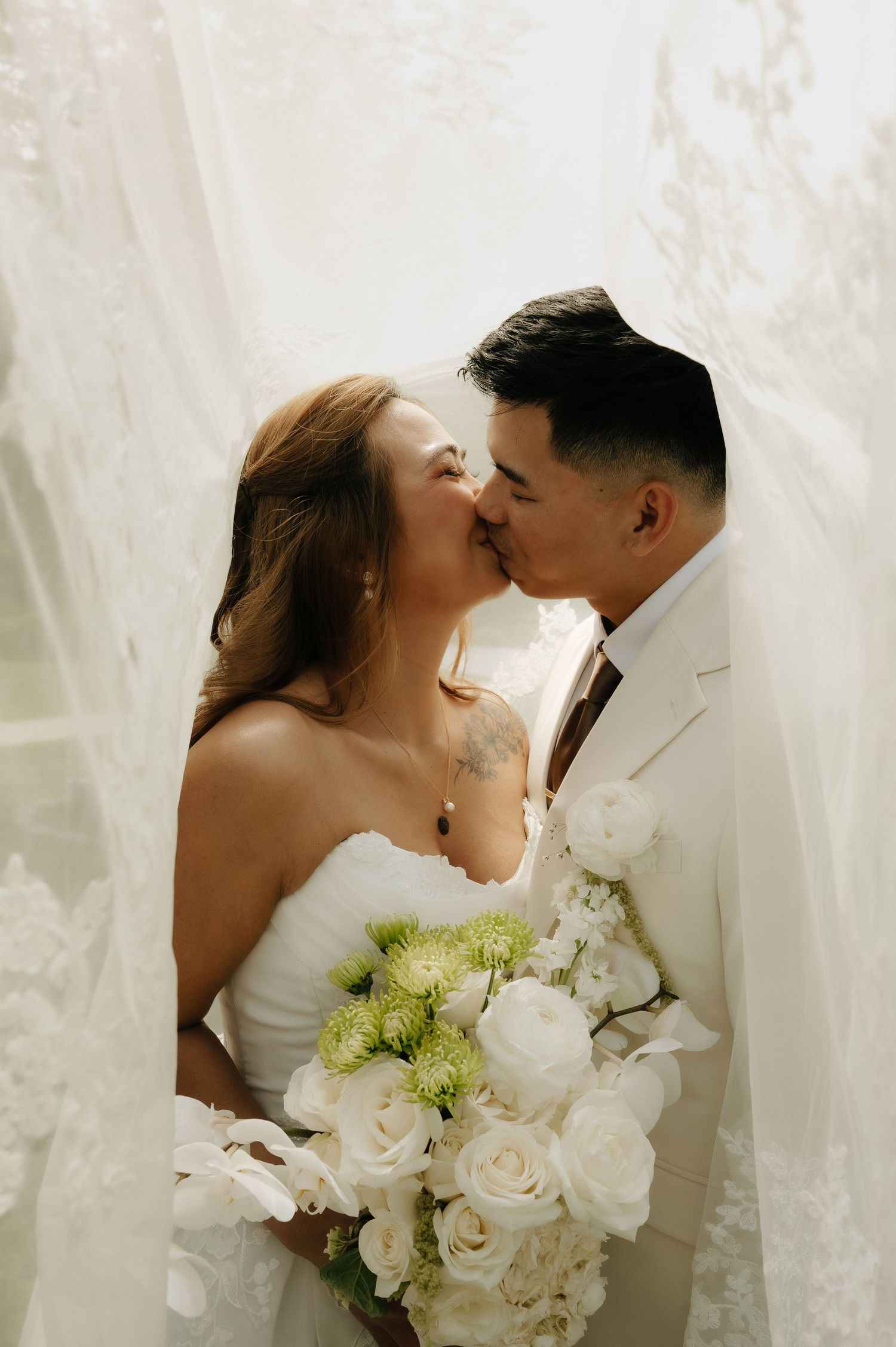 Bride and groom kissing between flowing white veils, bride holding bouquet of white roses and green flowers.