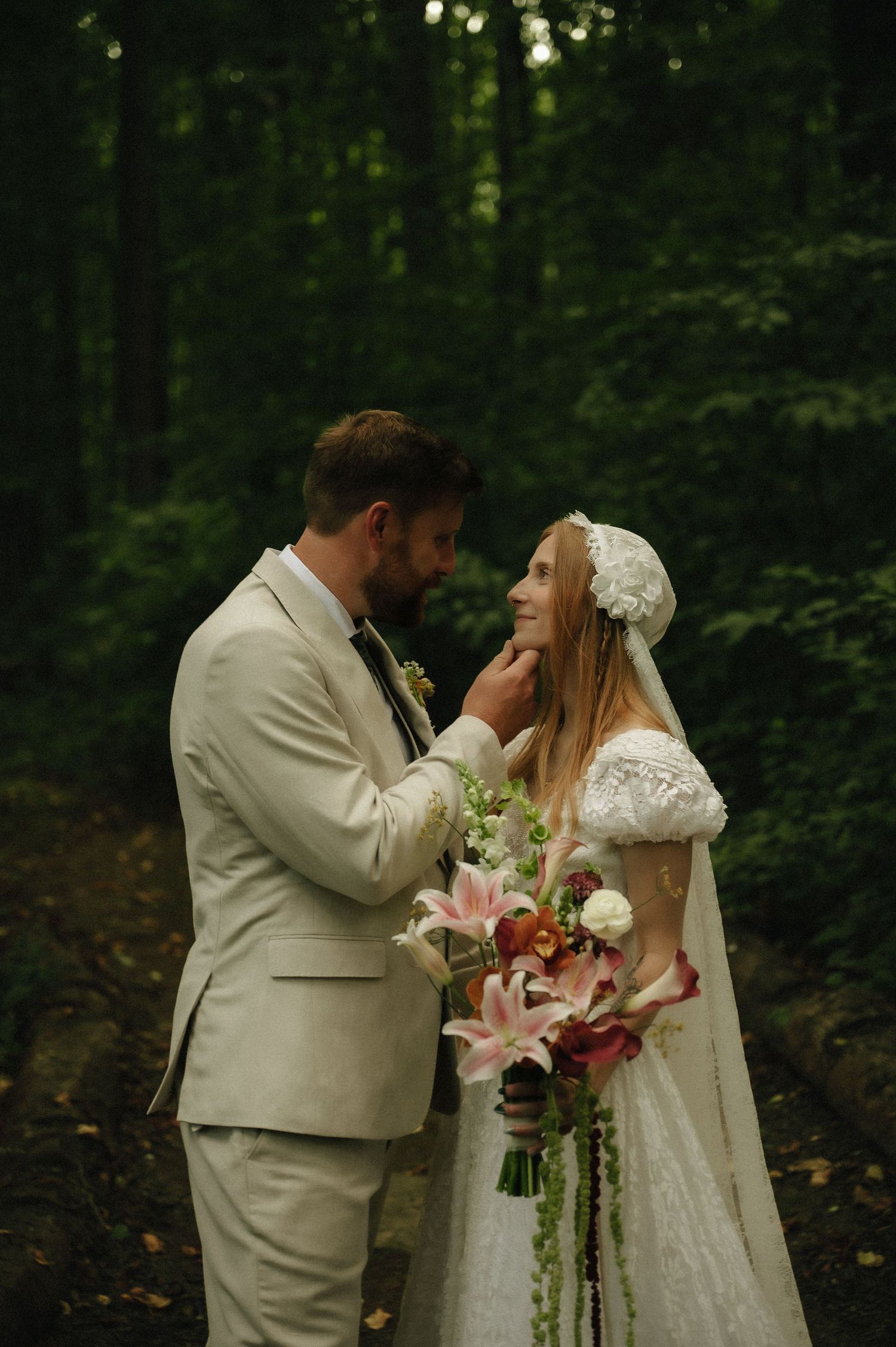 Bride and groom share a tender moment in a lush forest, she holds a pink lily bouquet wearing a lace dress and bonnet.