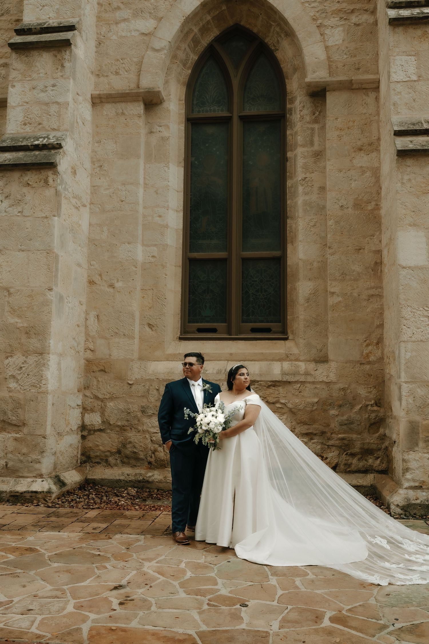 Wedding couple poses in front of gothic church window with flowing veil.