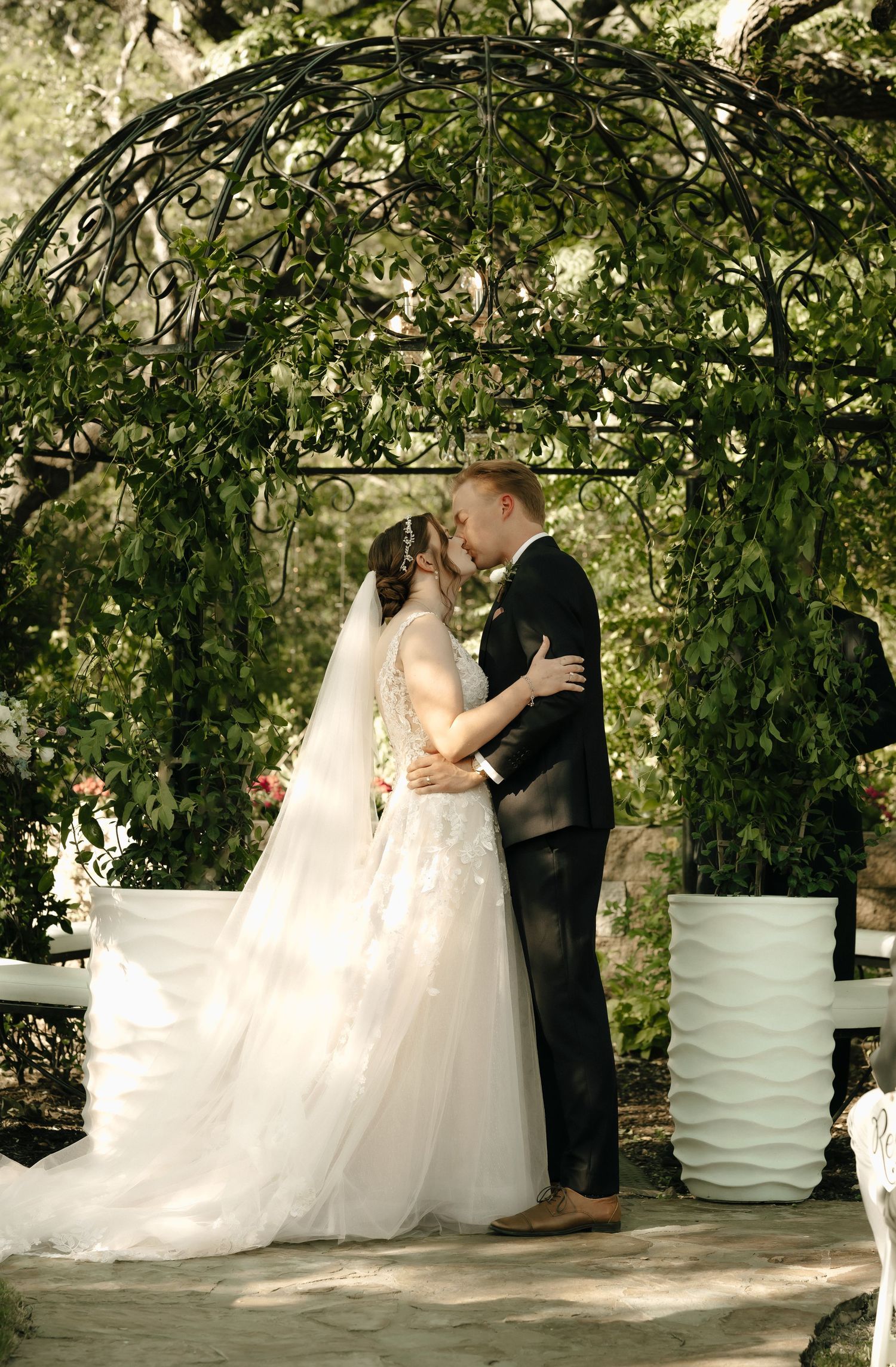 Newlyweds share intimate moment under green garden archway with white planters.