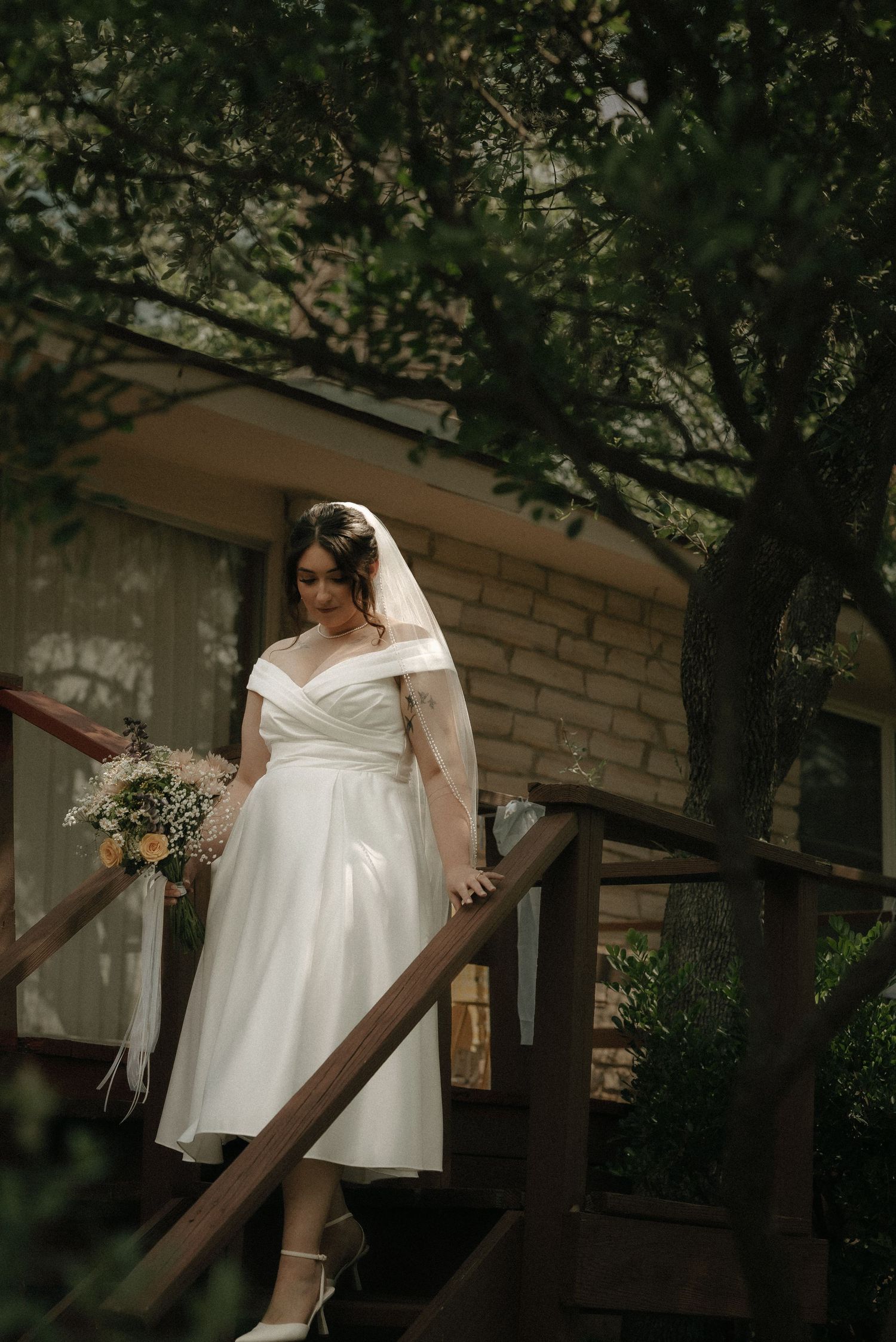 A bride in a tea-length dress and veil stands on wooden stairs surrounded by greenery.