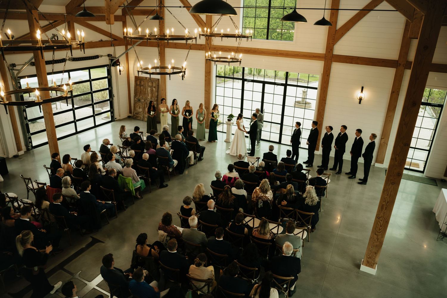 Aerial view of an elegant indoor wedding ceremony in a rustic barn venue with wood beams, chandeliers, and large windows.