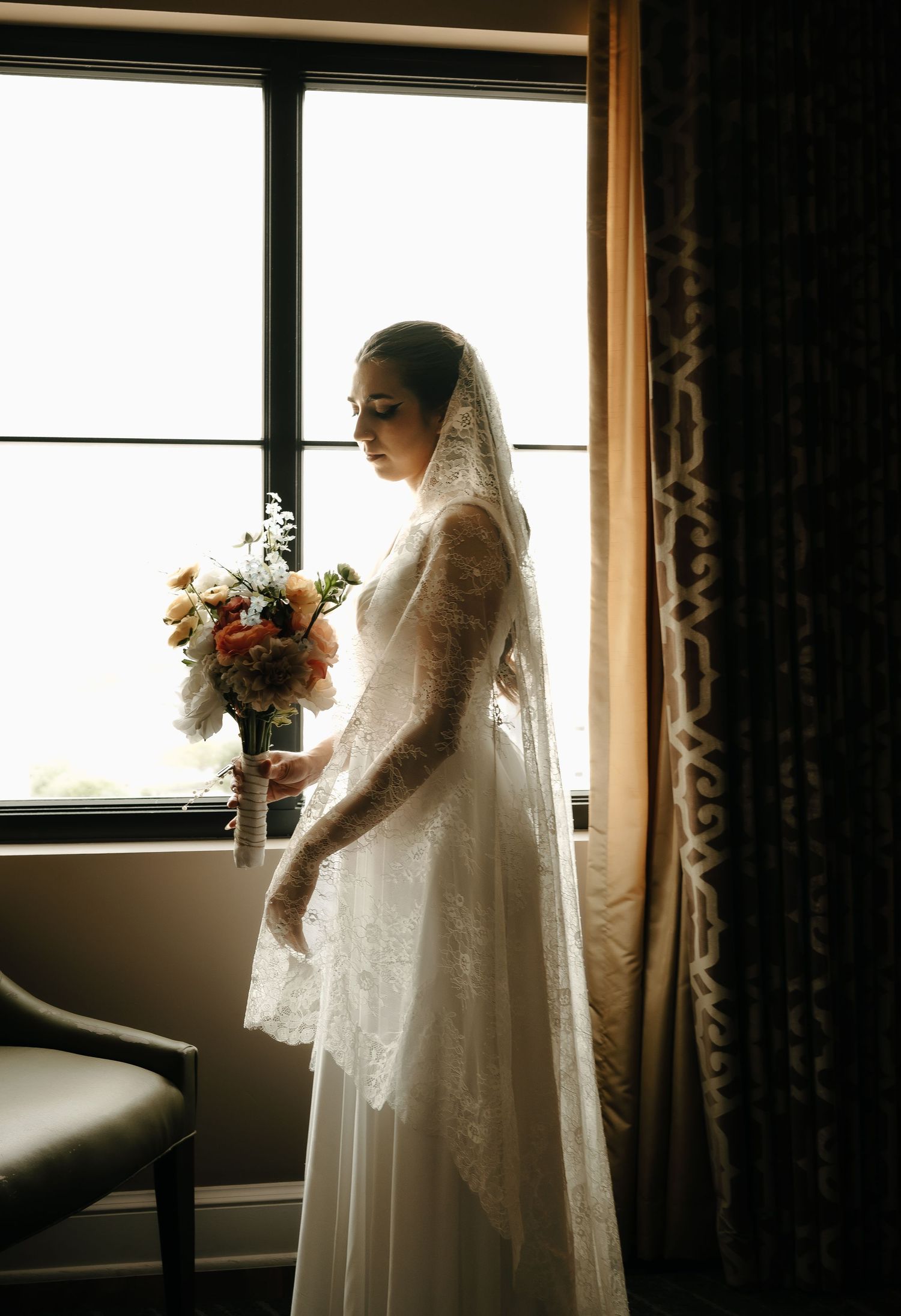 Silhouetted bride in lace gown holds bouquet by window in dramatic lighting.