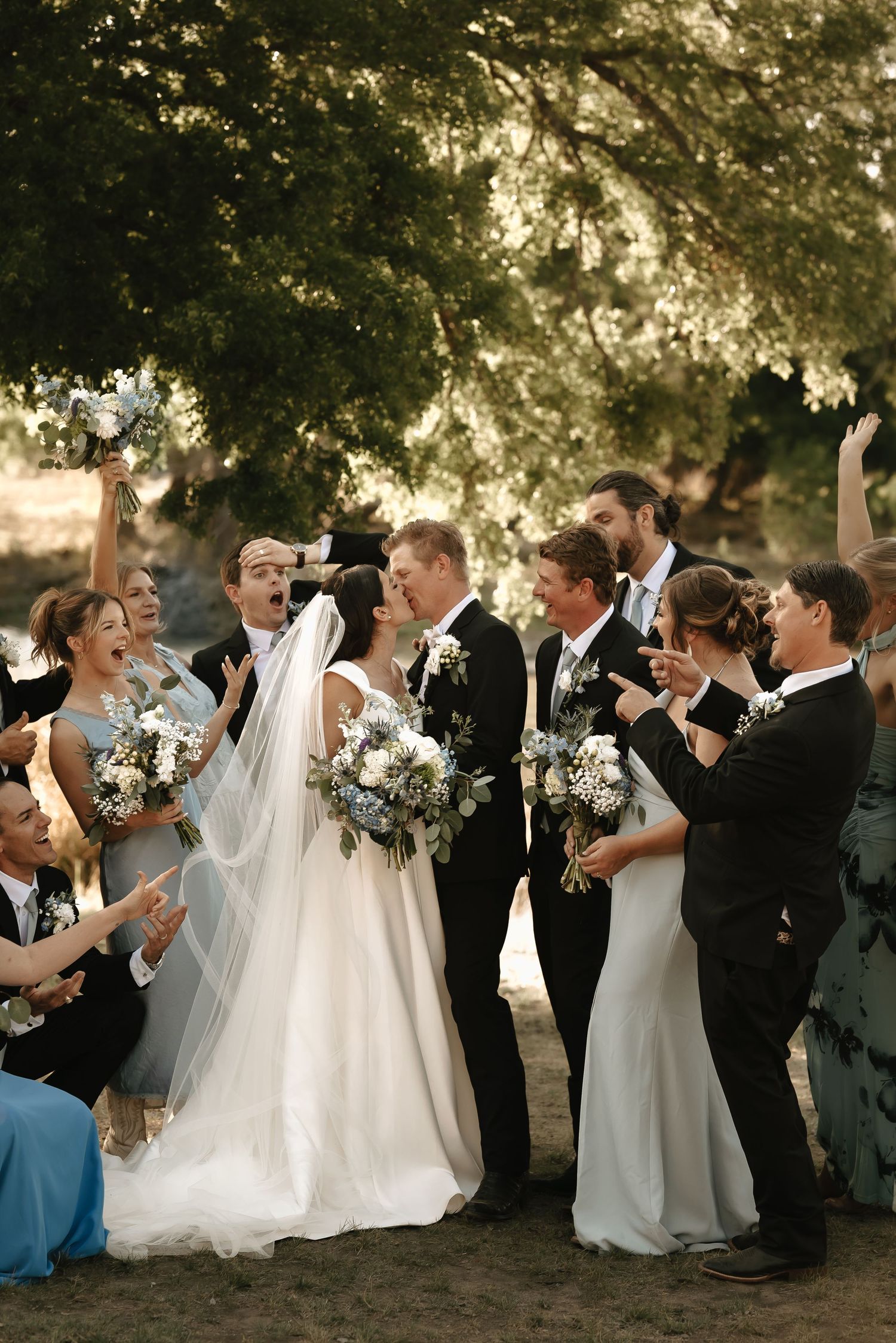 Wedding party celebrates outdoors under trees with joyful expressions.
