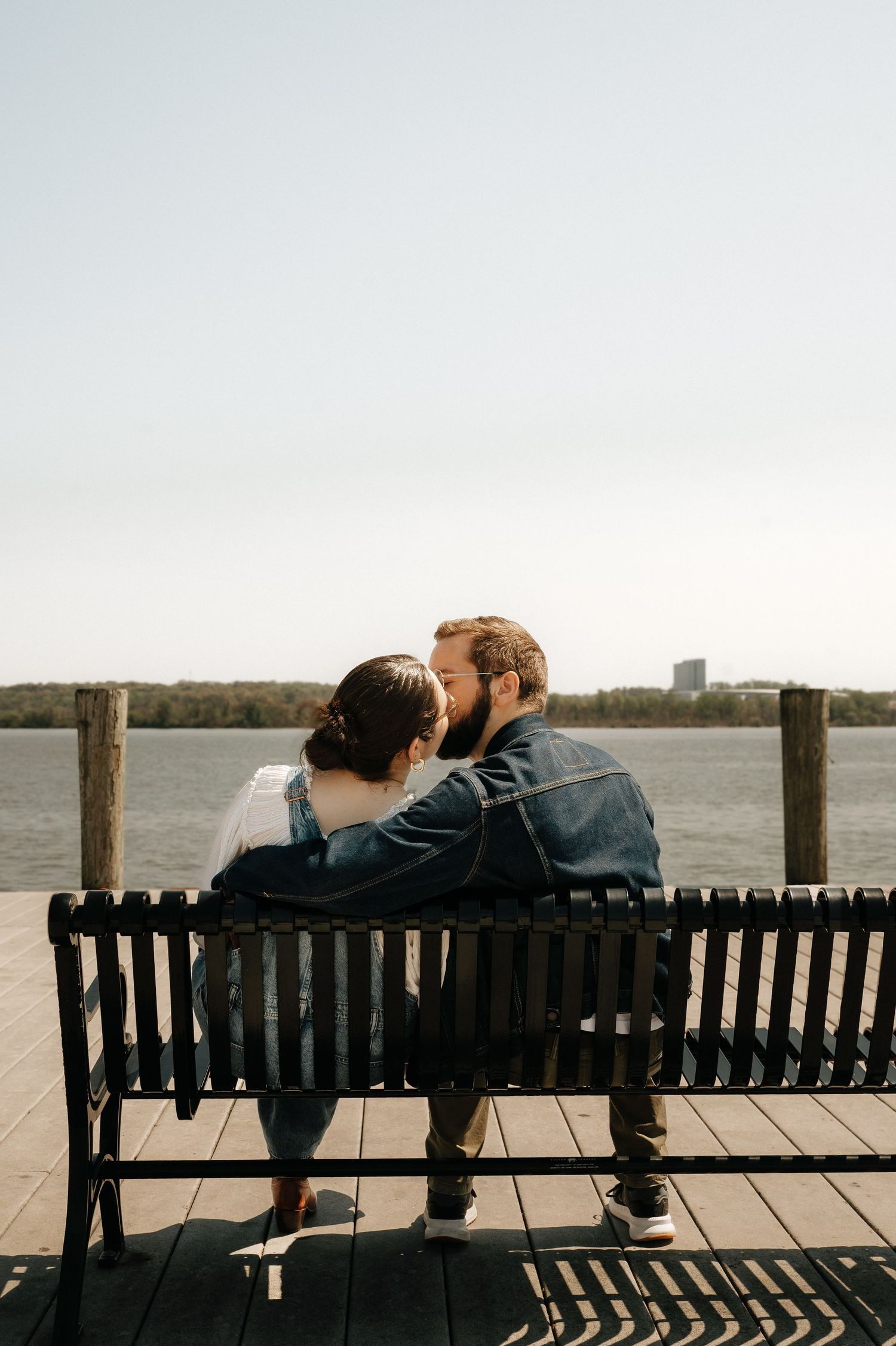 Couple sharing a romantic moment on a waterfront bench, with a serene river and clear sky backdrop.
