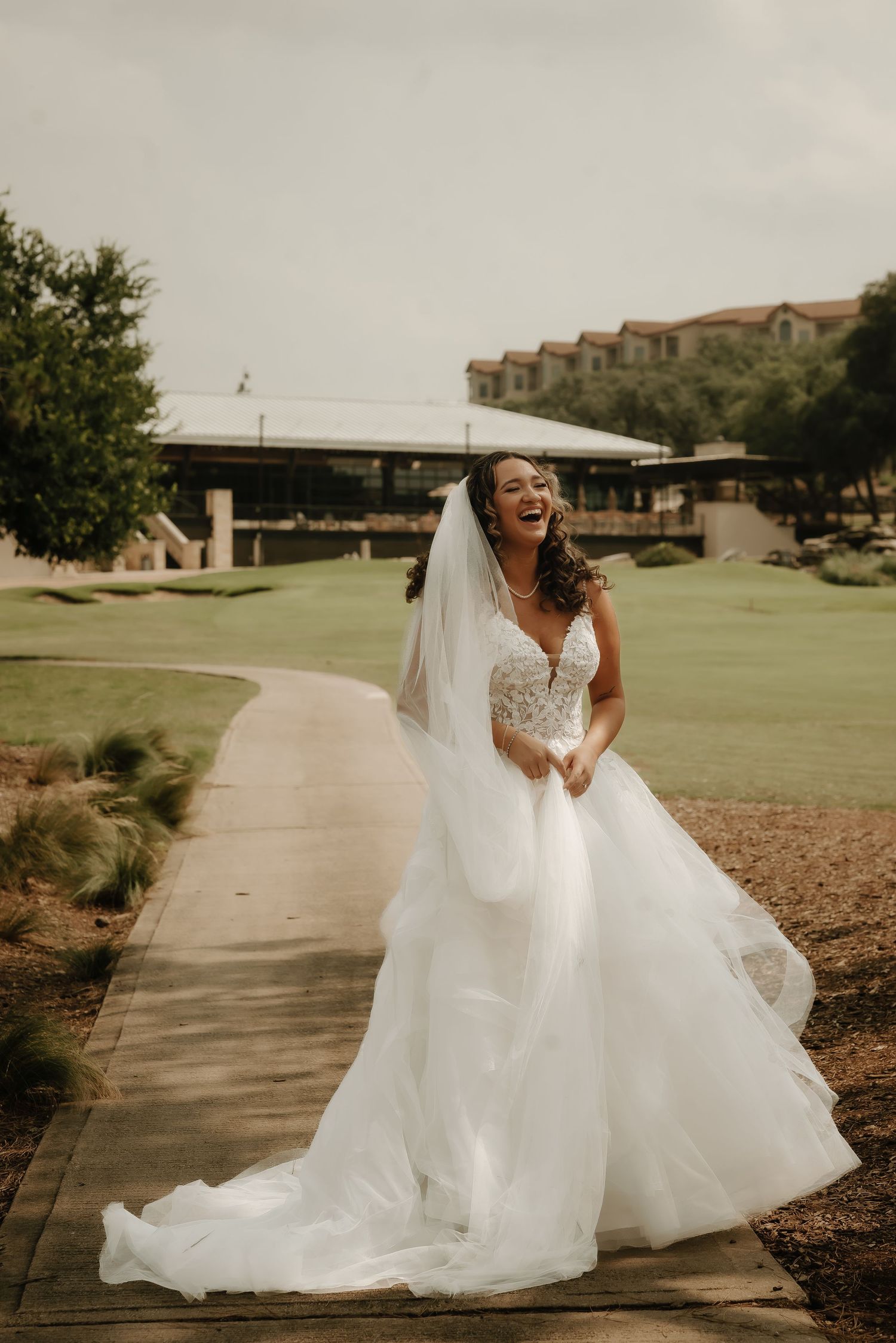 A bride in a flowing white gown twirls on a pathway surrounded by manicured grounds.