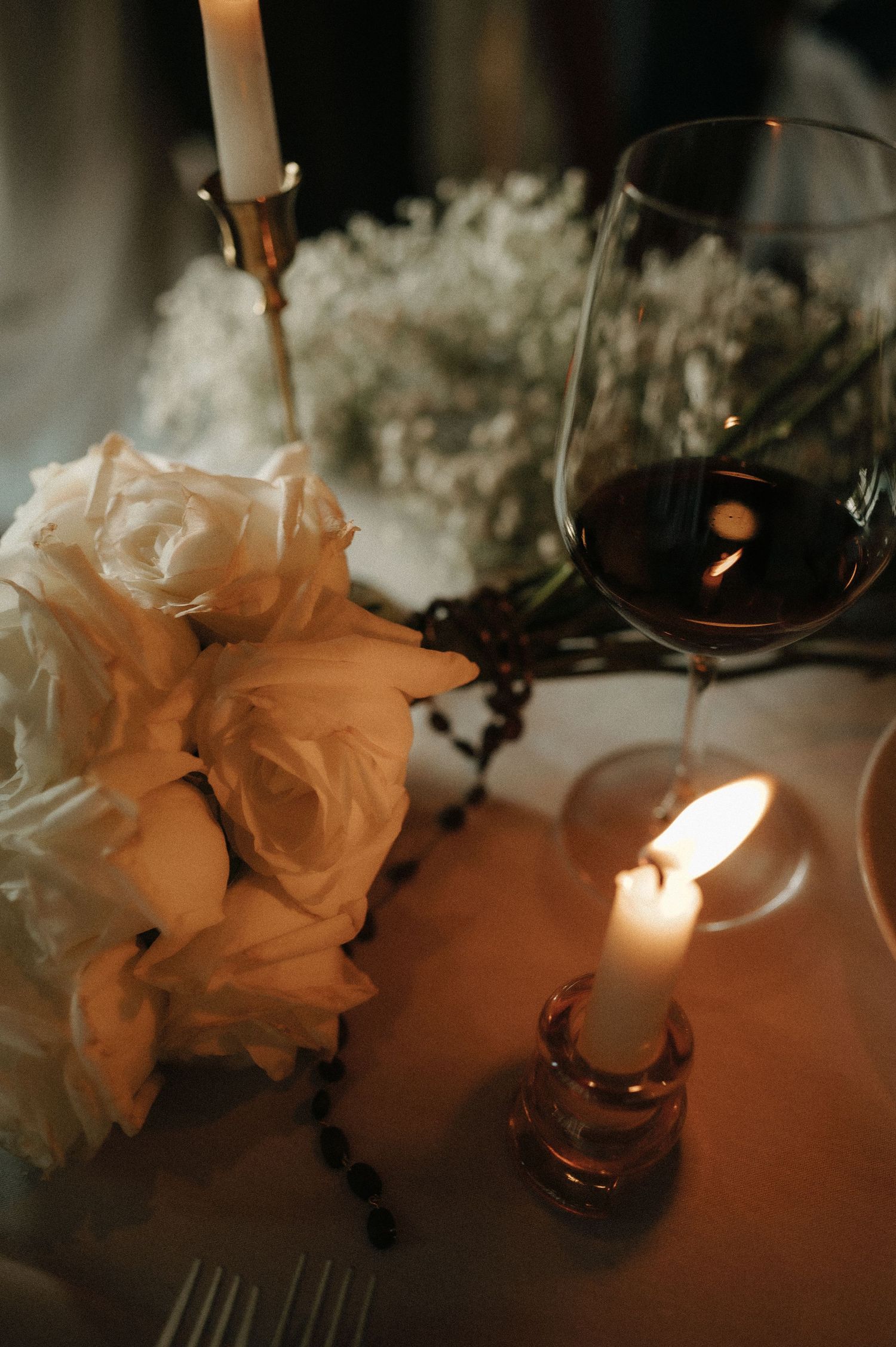 A romantic table setting with lit candles, white roses, and red wine in ambient lighting.