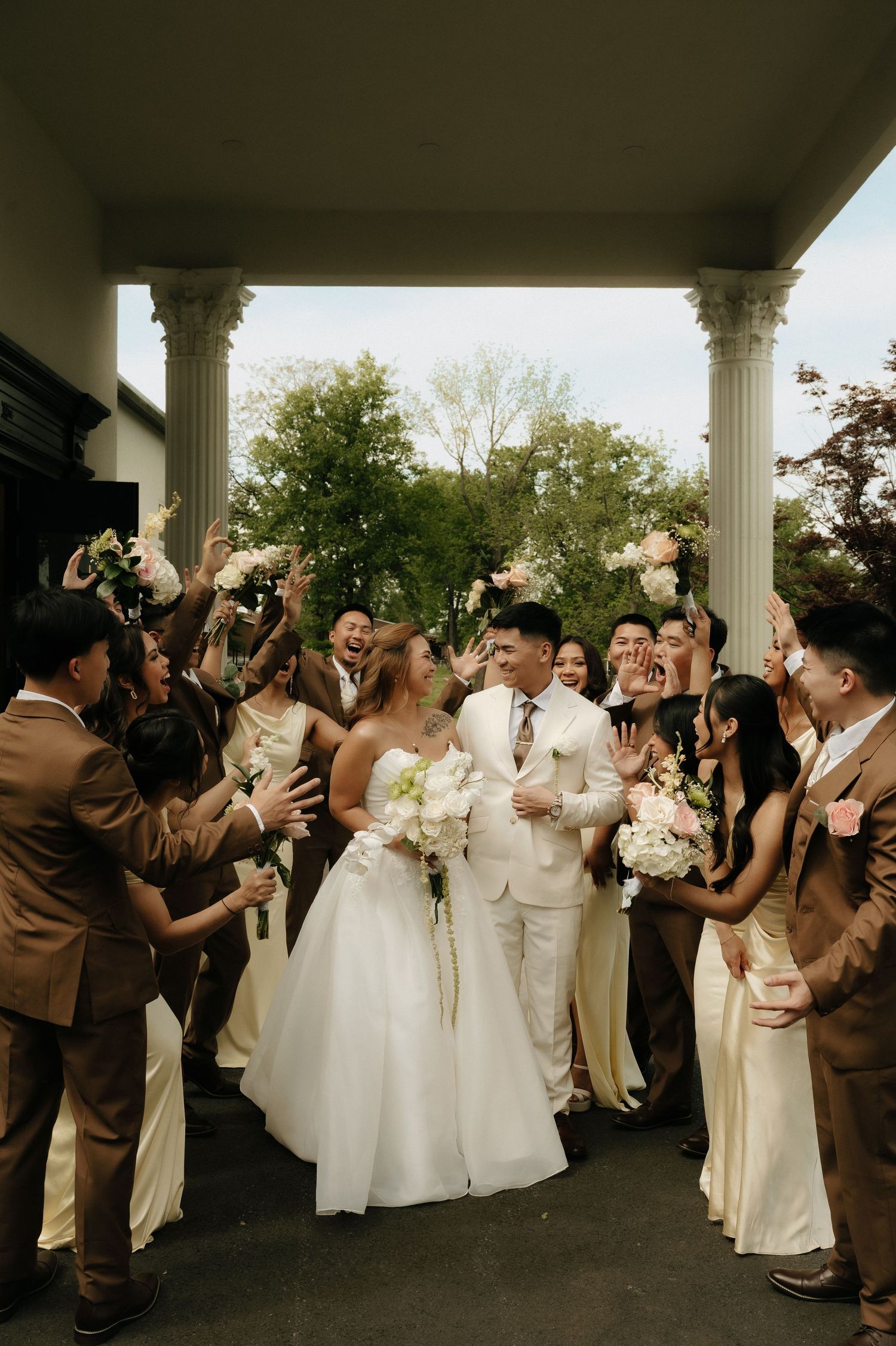 Bride and groom walk through cheering wedding party under white columns, surrounded by bouquets and tan bridesmaid dresses.