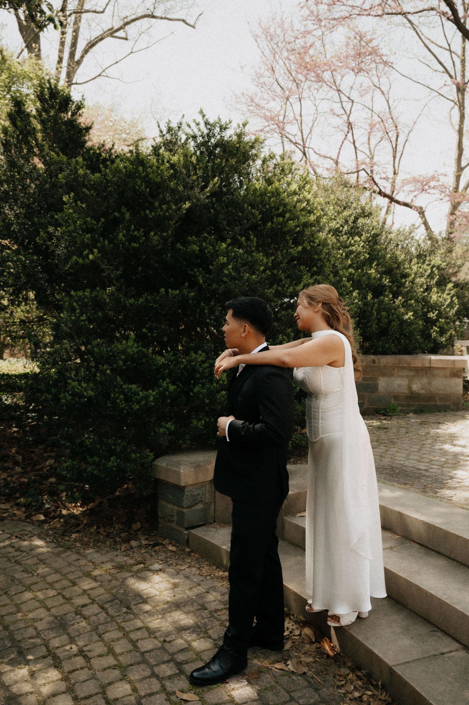 Bride in white dress adjusts groom's suit jacket outdoors near lush green hedges on cobblestone steps.