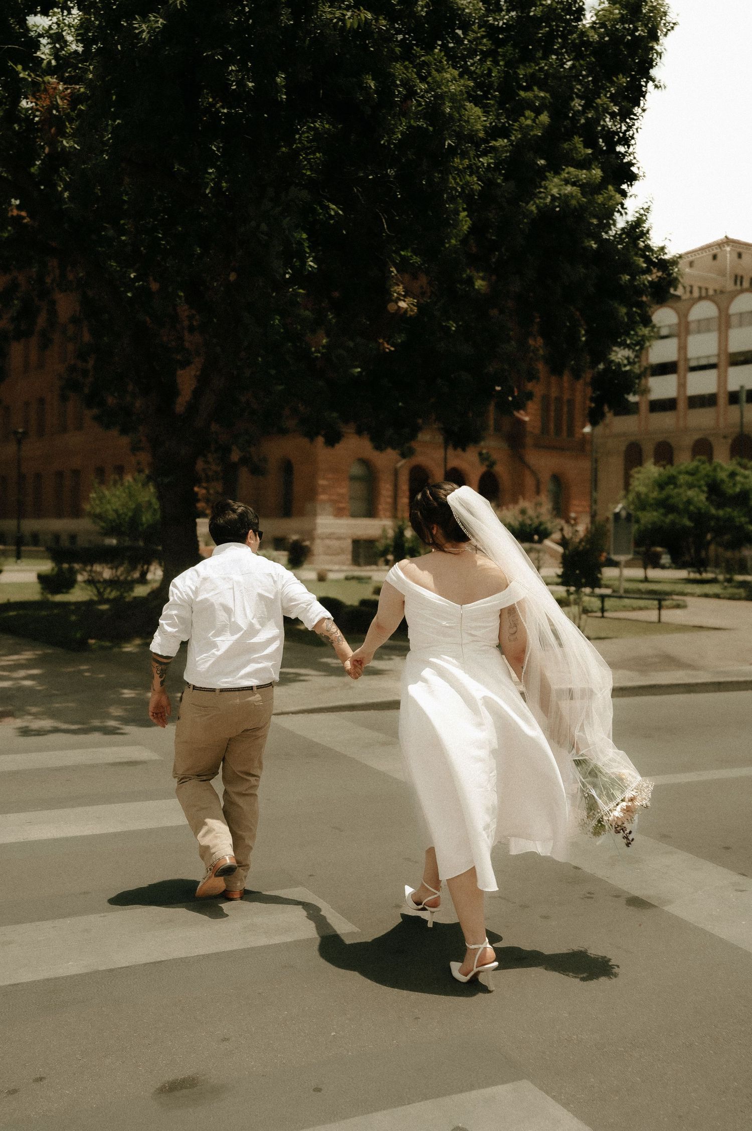 A newlywed couple walking hand in hand across a city street in vintage-inspired wedding attire.