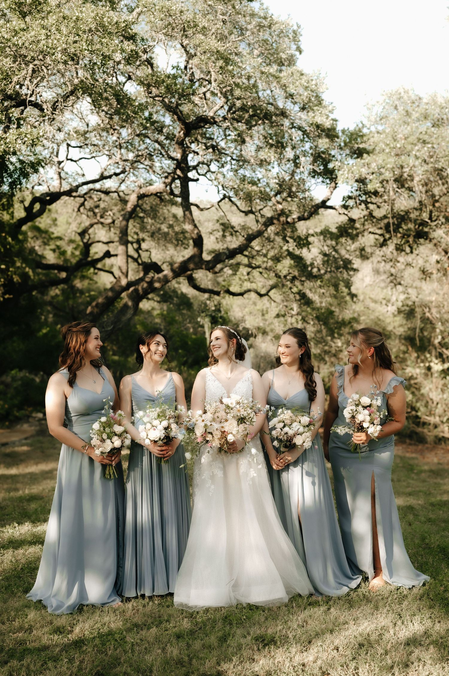 Wedding party in dusty blue dresses and white gown posed on hillside with floral bouquets.