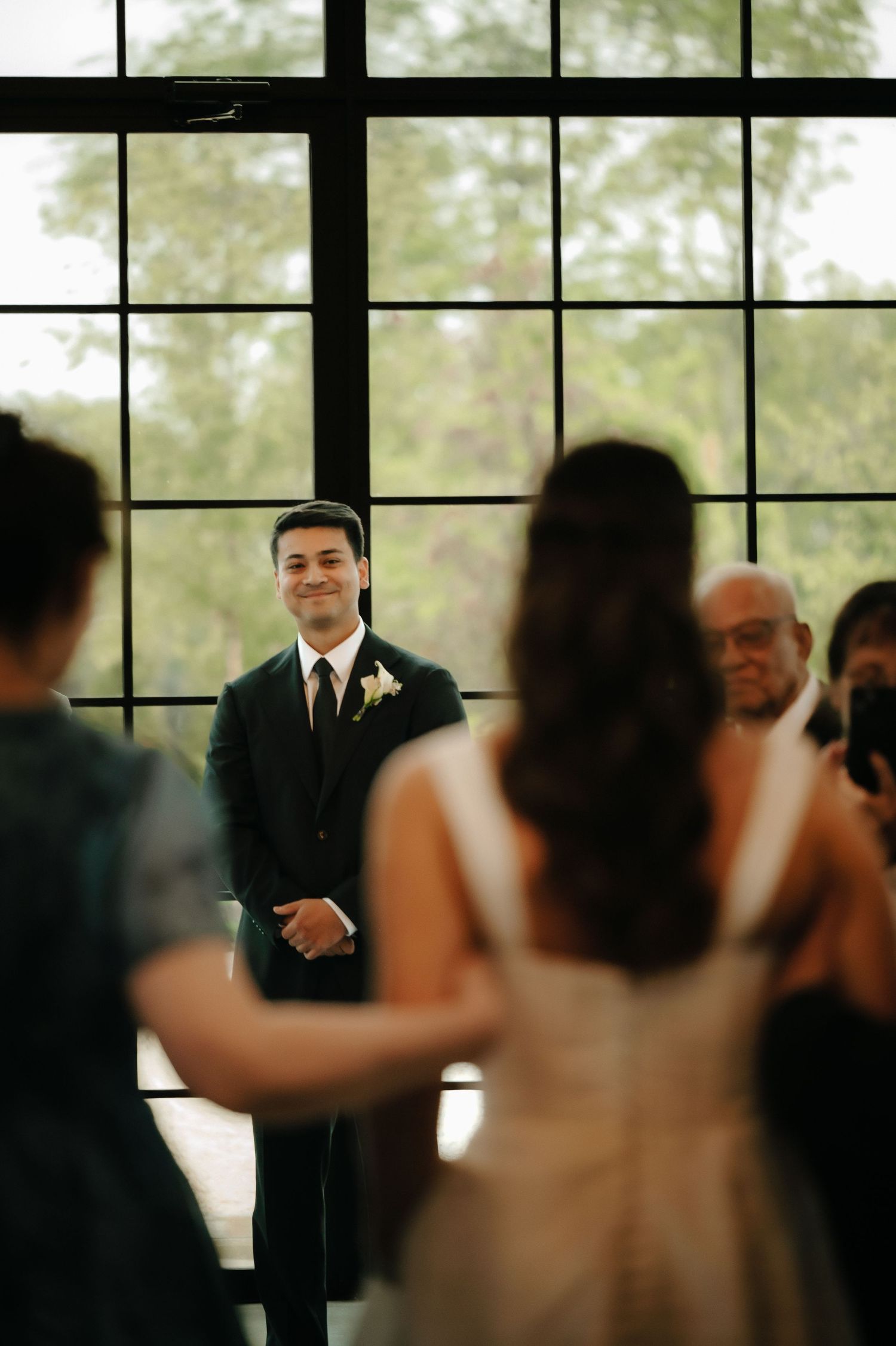 Groom smiling at bride walking down the aisle during indoor wedding ceremony with large windows.