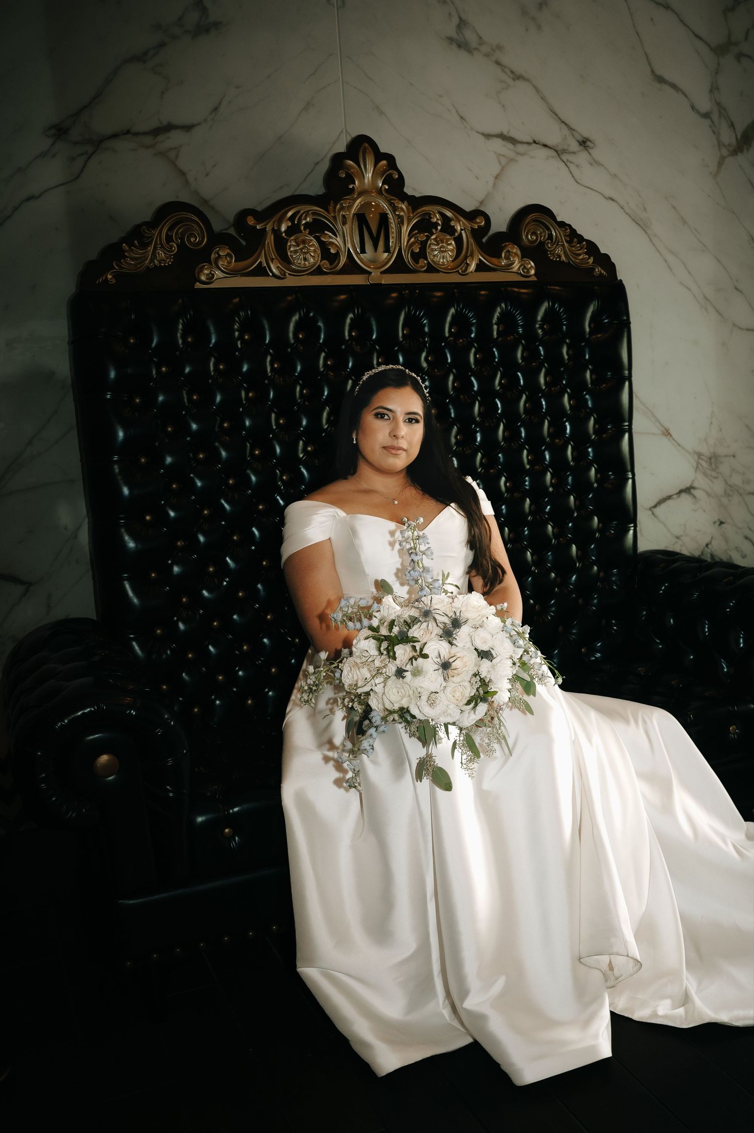 Bride seated on ornate black throne chair holding white floral bouquet.