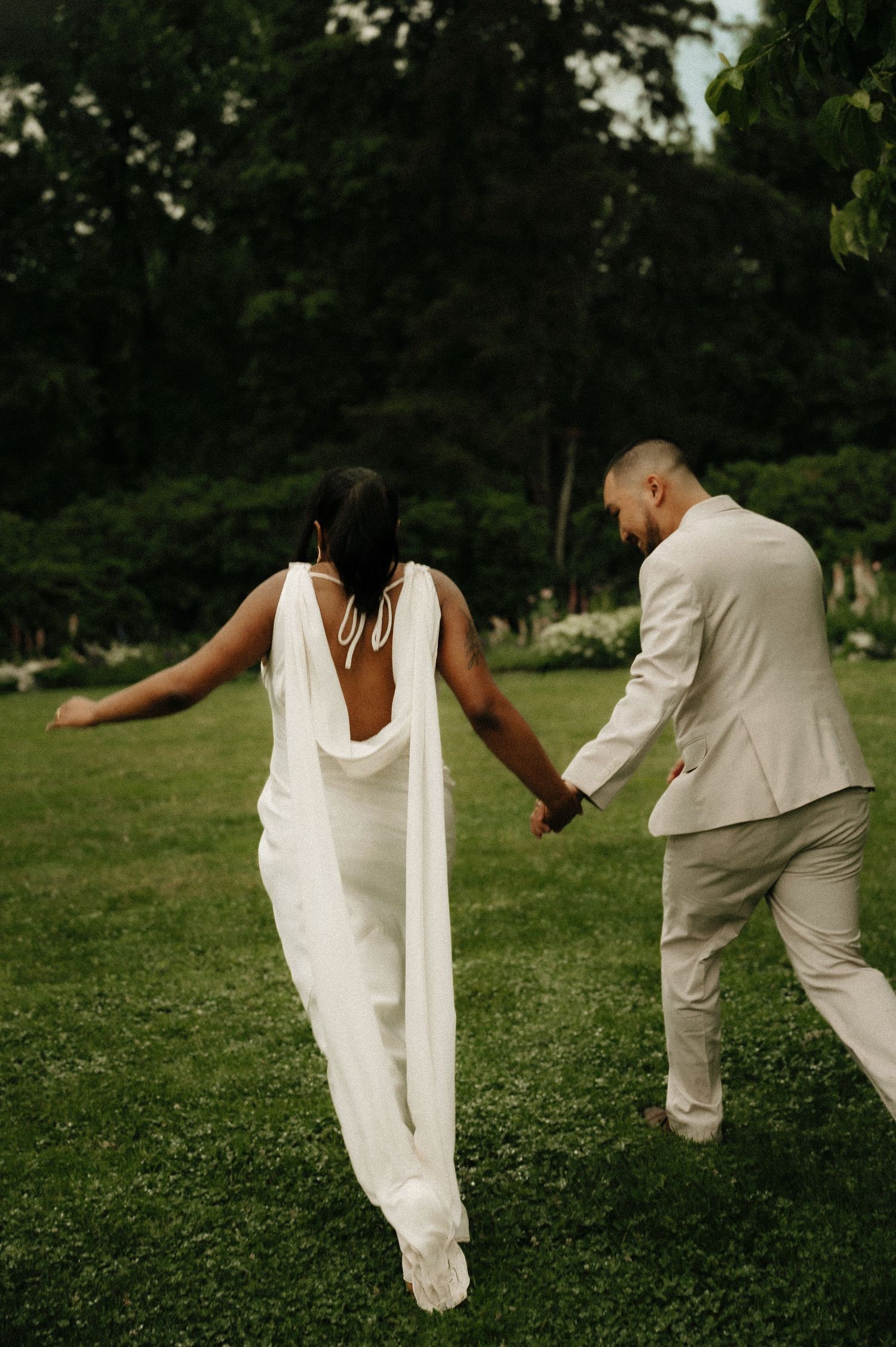 A bride in a white backless gown and groom in a cream suit hold hands while running across a lush green lawn.