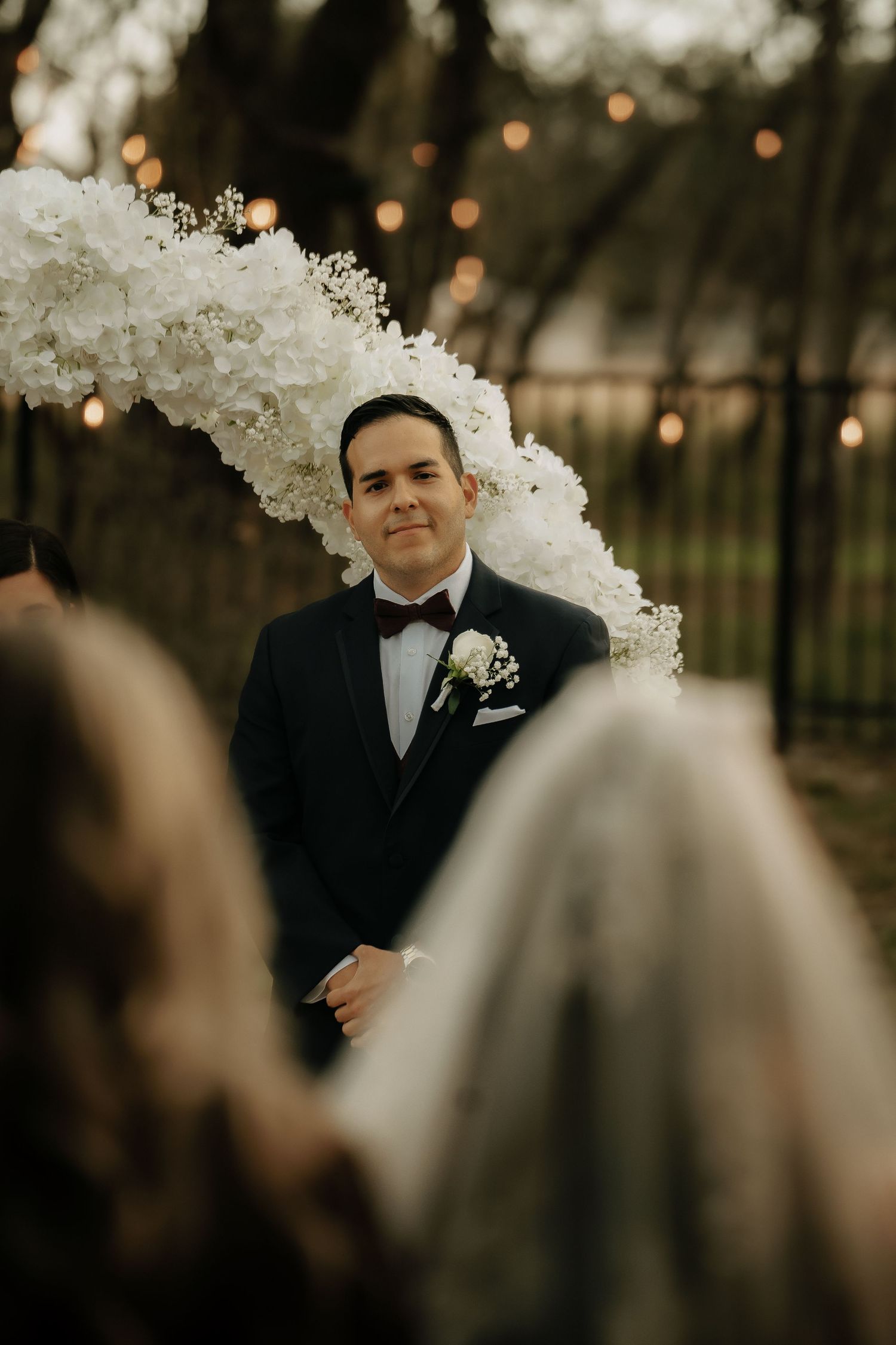 Groom in classic tuxedo stands under an elegant white floral arch during outdoor ceremony.