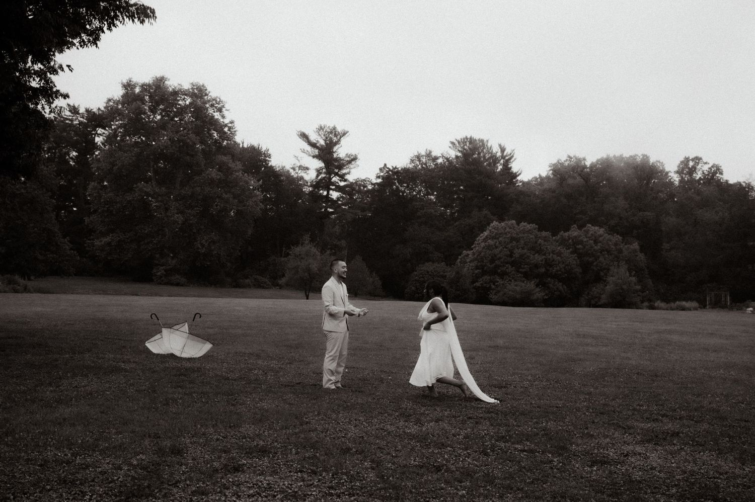 Black and white photo of wedding couple in open field with flowing bridal veil caught in breeze.