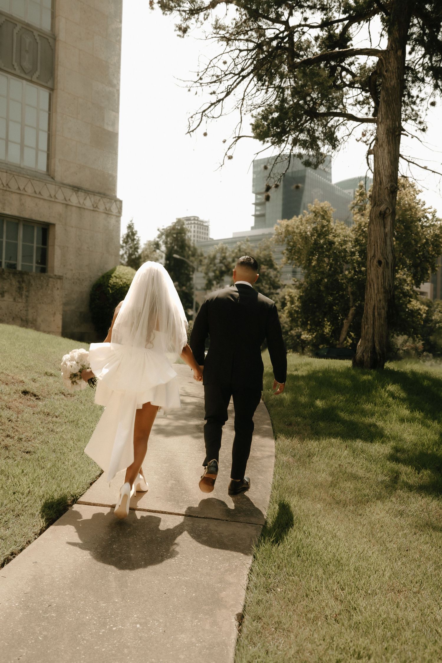 A bride and groom walk together on a garden path outside a stone building on their wedding day.