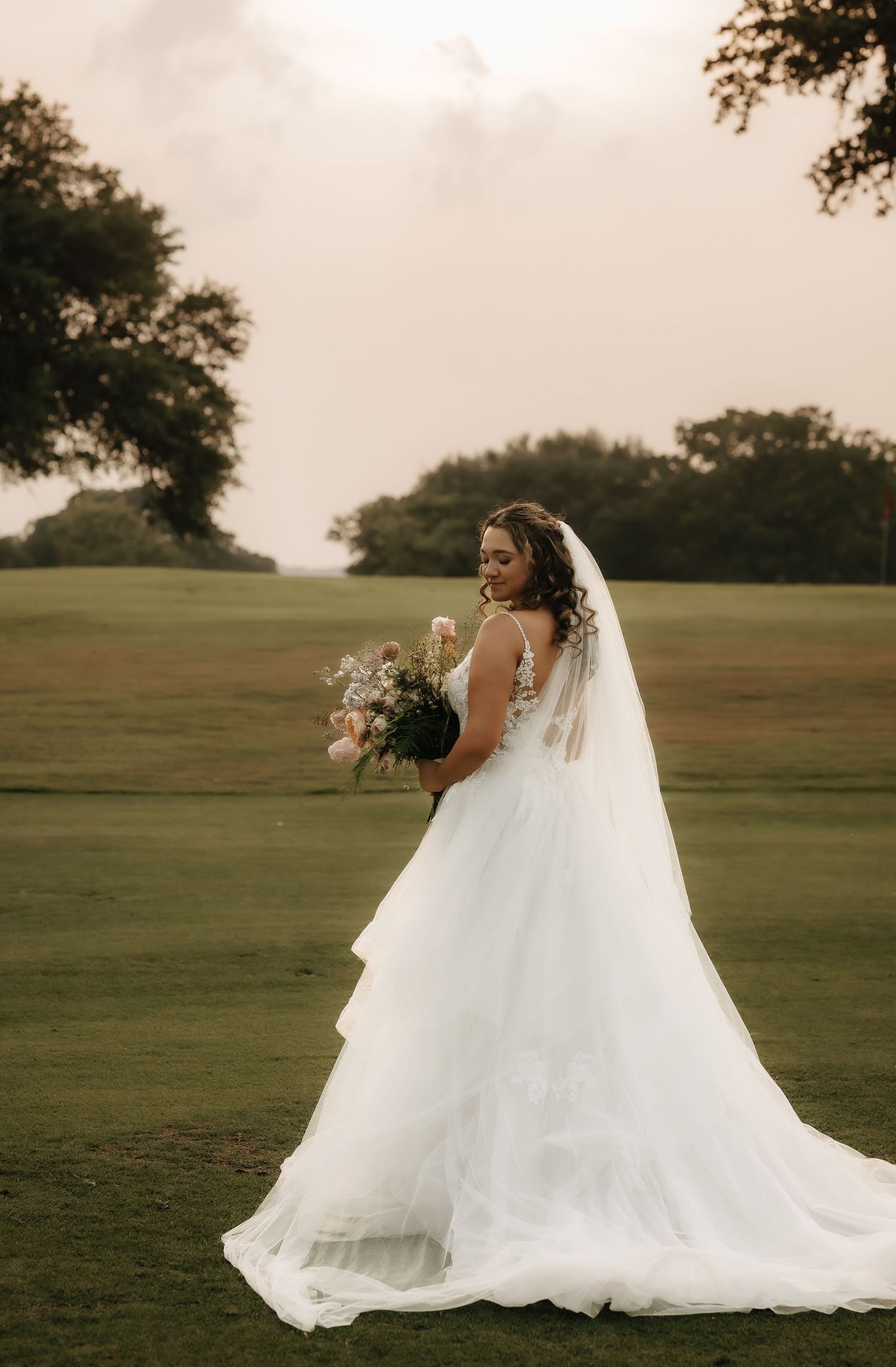 A bride in a flowing white wedding gown poses gracefully in an open field during golden hour.