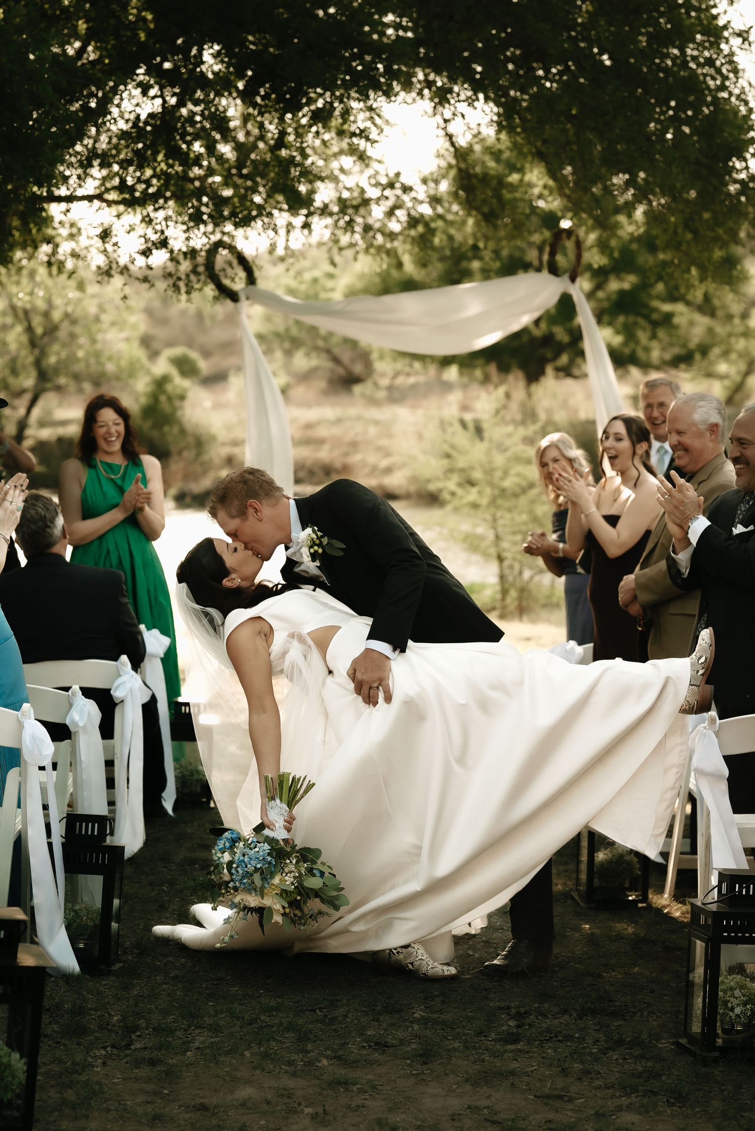 Wedding party posing dramatically on white chairs under trees during outdoor ceremony.