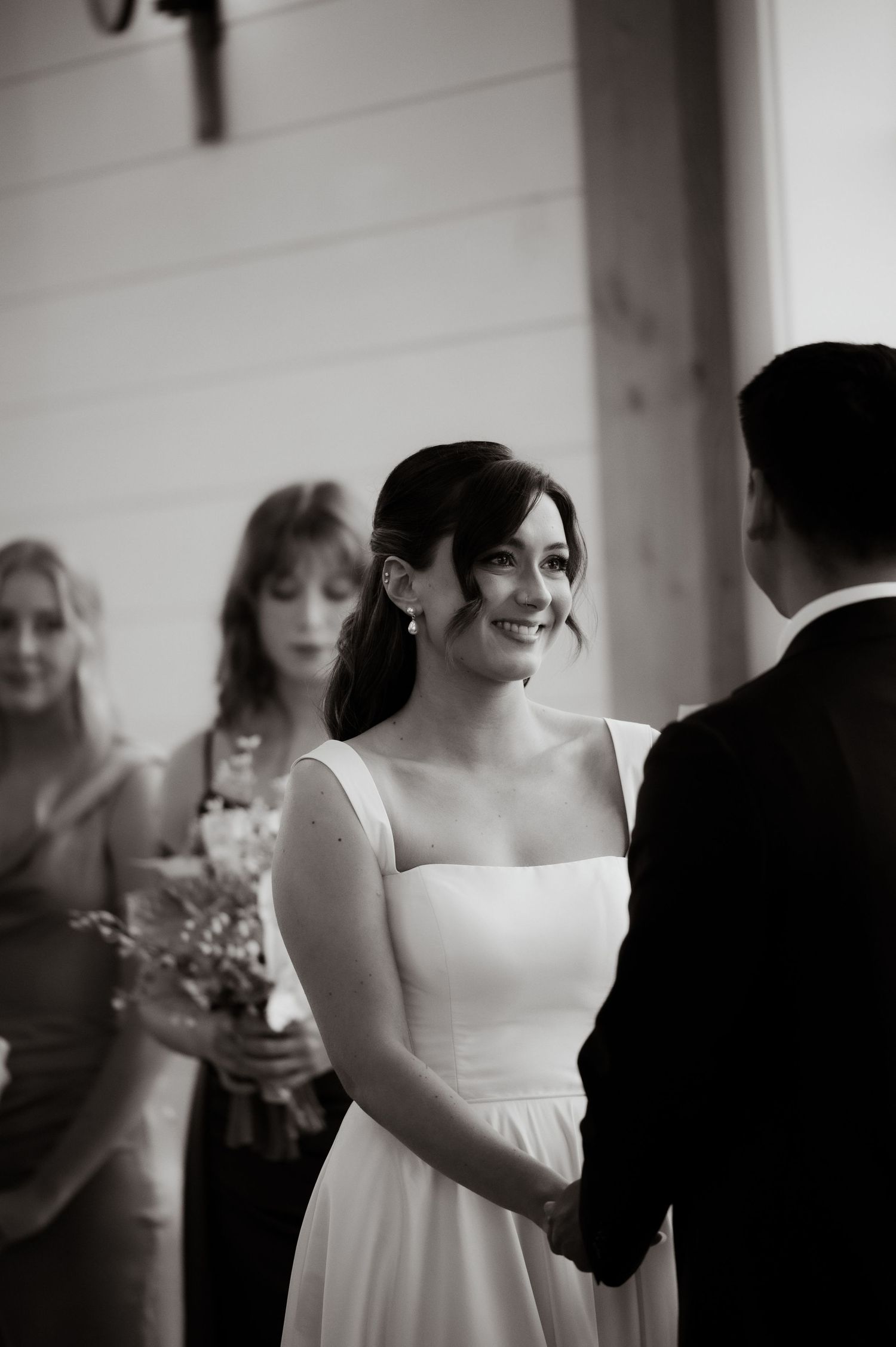 Black and white photo of a smiling bride in a white gown holding hands with groom during wedding ceremony.
