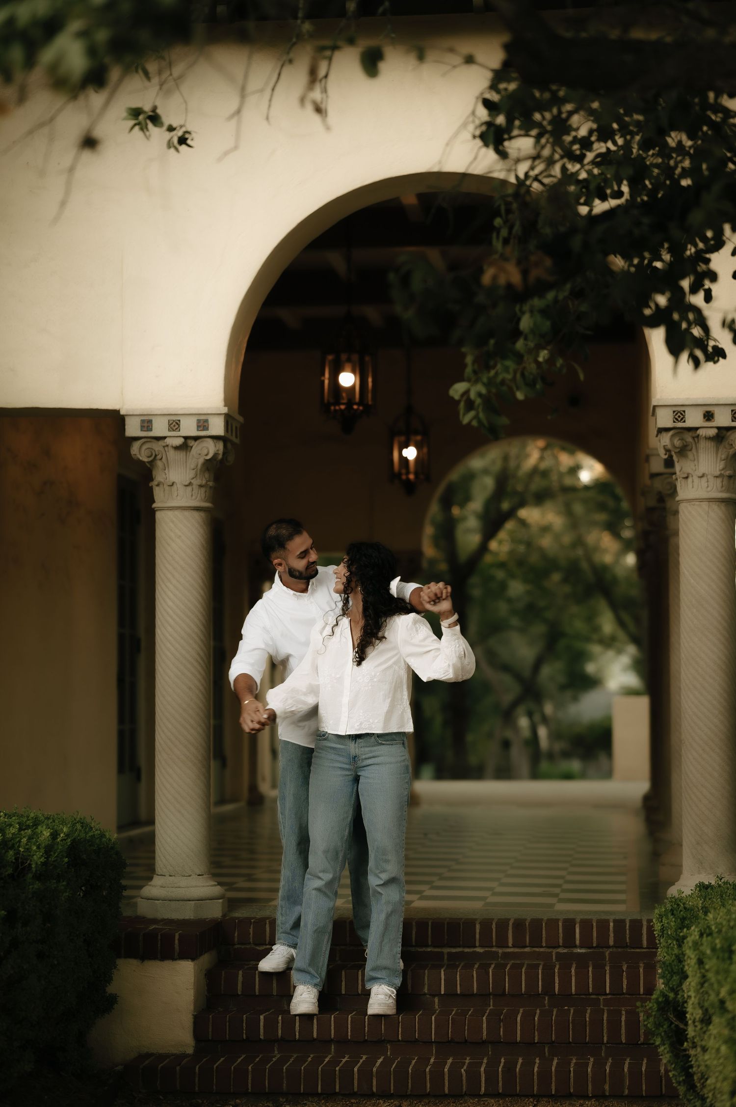 Playful couple on brick steps under arched colonnade in matching white tops.