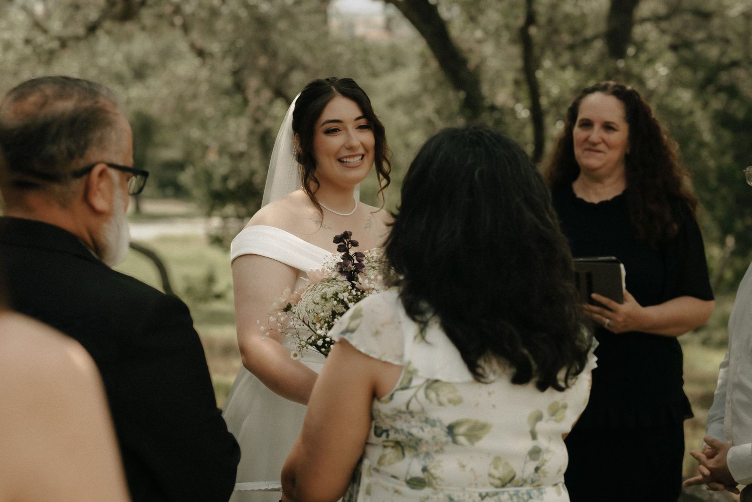 Wedding guests gather outdoors under trees for an intimate ceremony.