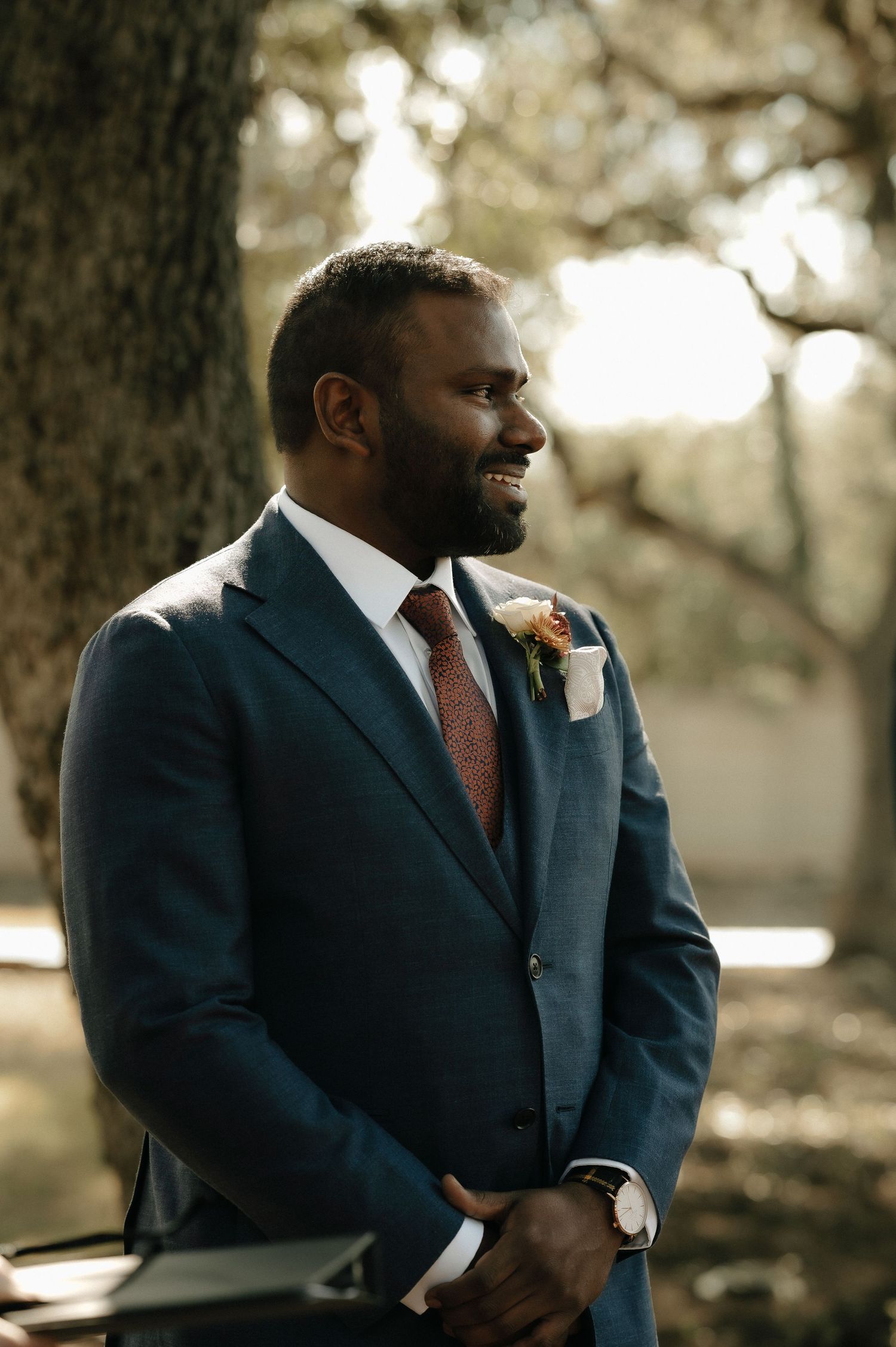 A groomsman in a navy suit and coral tie smiles during an outdoor wedding.
