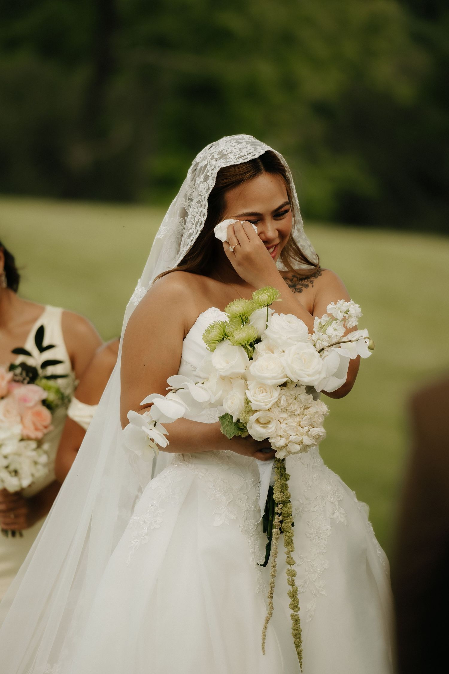Emotional bride crying while holding white bouquet of roses and hydrangeas, wearing lace veil at outdoor wedding ceremony.