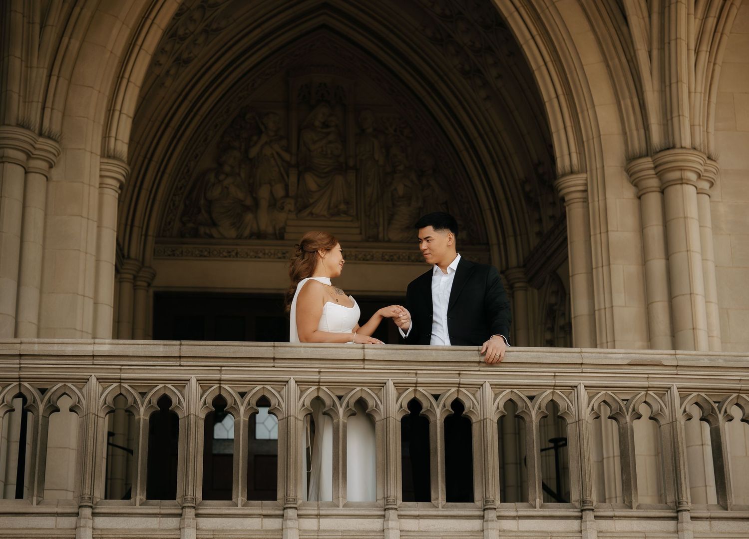 Bride and groom share a romantic moment on a Gothic cathedral balcony, dressed in elegant wedding attire.