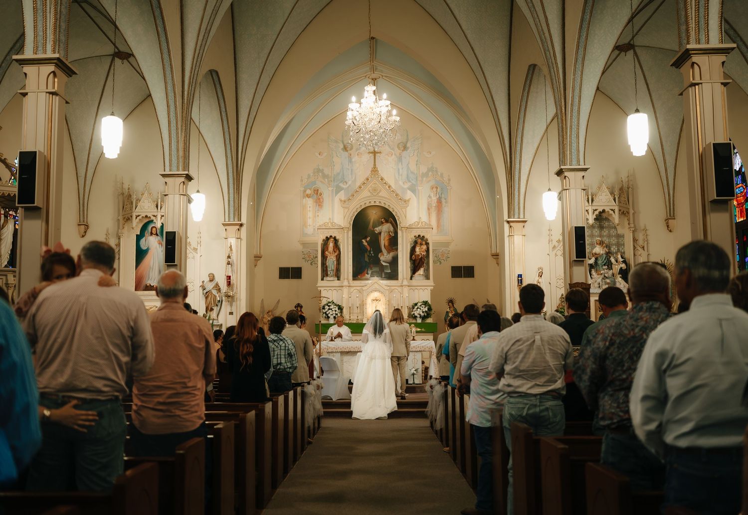 Interior of a cathedral-style church with vaulted ceilings and chandeliers during a wedding ceremony.