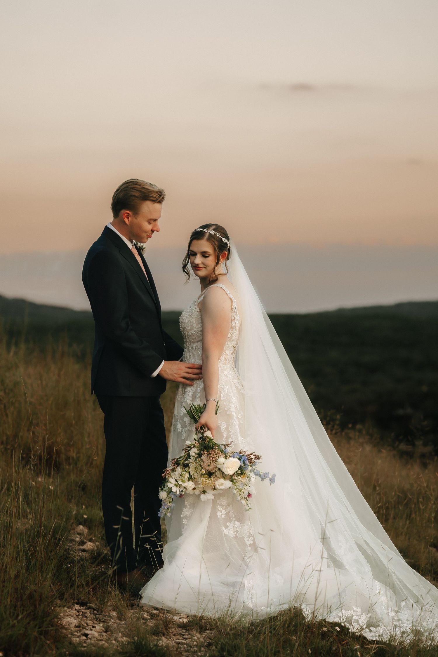 Wedding couple embracing at sunset in flowing white gown with dramatic mountain backdrop.