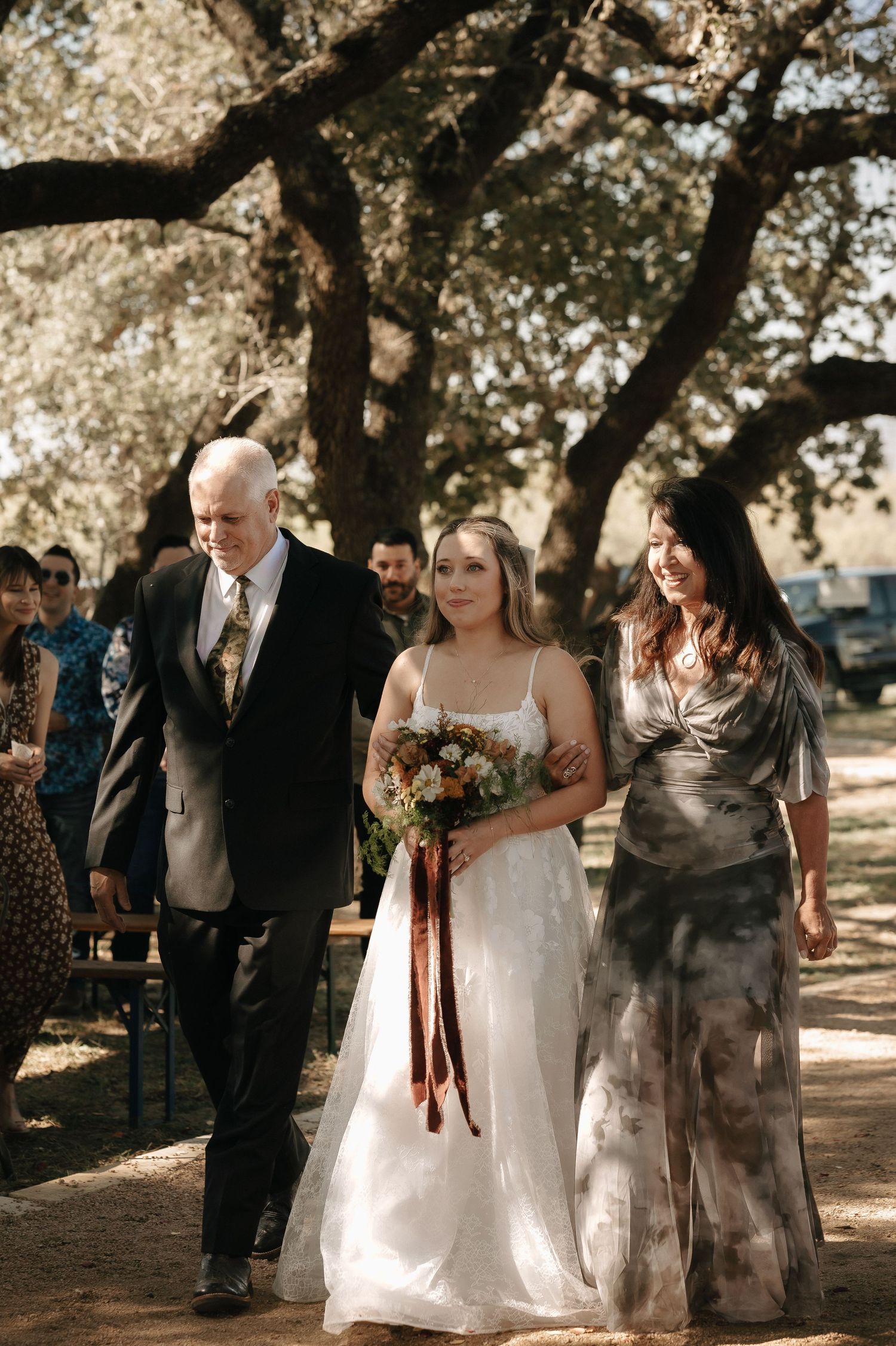 A wedding party walks together under trees during an outdoor ceremony with a beautiful white gown.