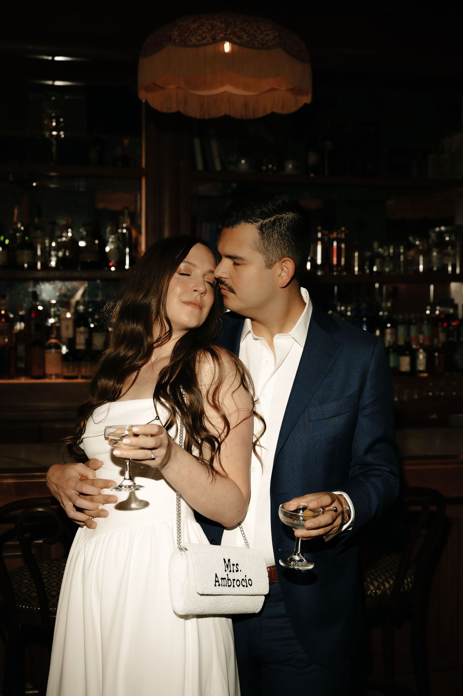 Couple in elegant attire sharing wine at a dark bar, woman in white dress holding Mrs. Ambrosio purse.