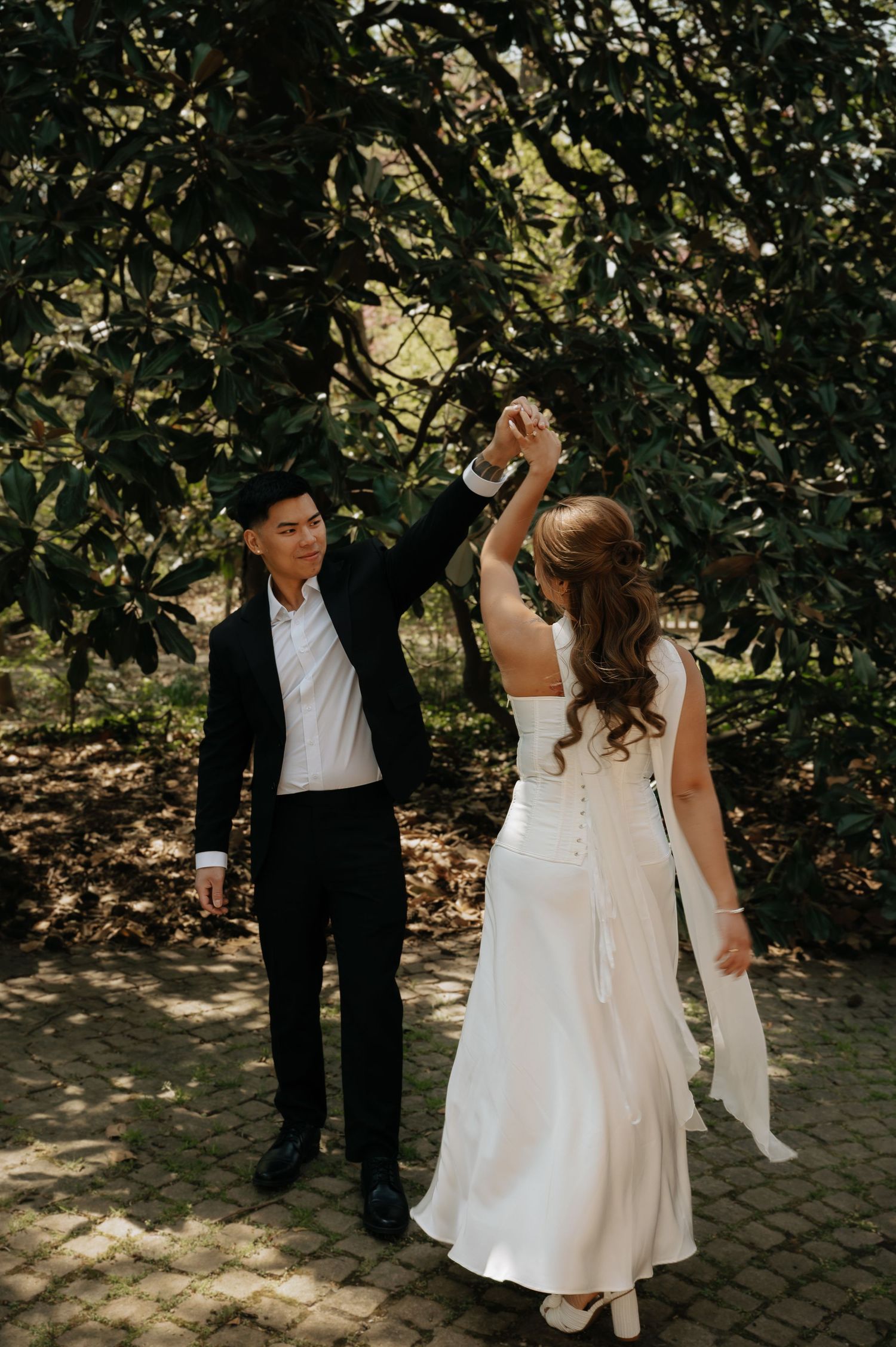 Bride in white gown being twirled by groom in black suit on brick path under lush green trees.