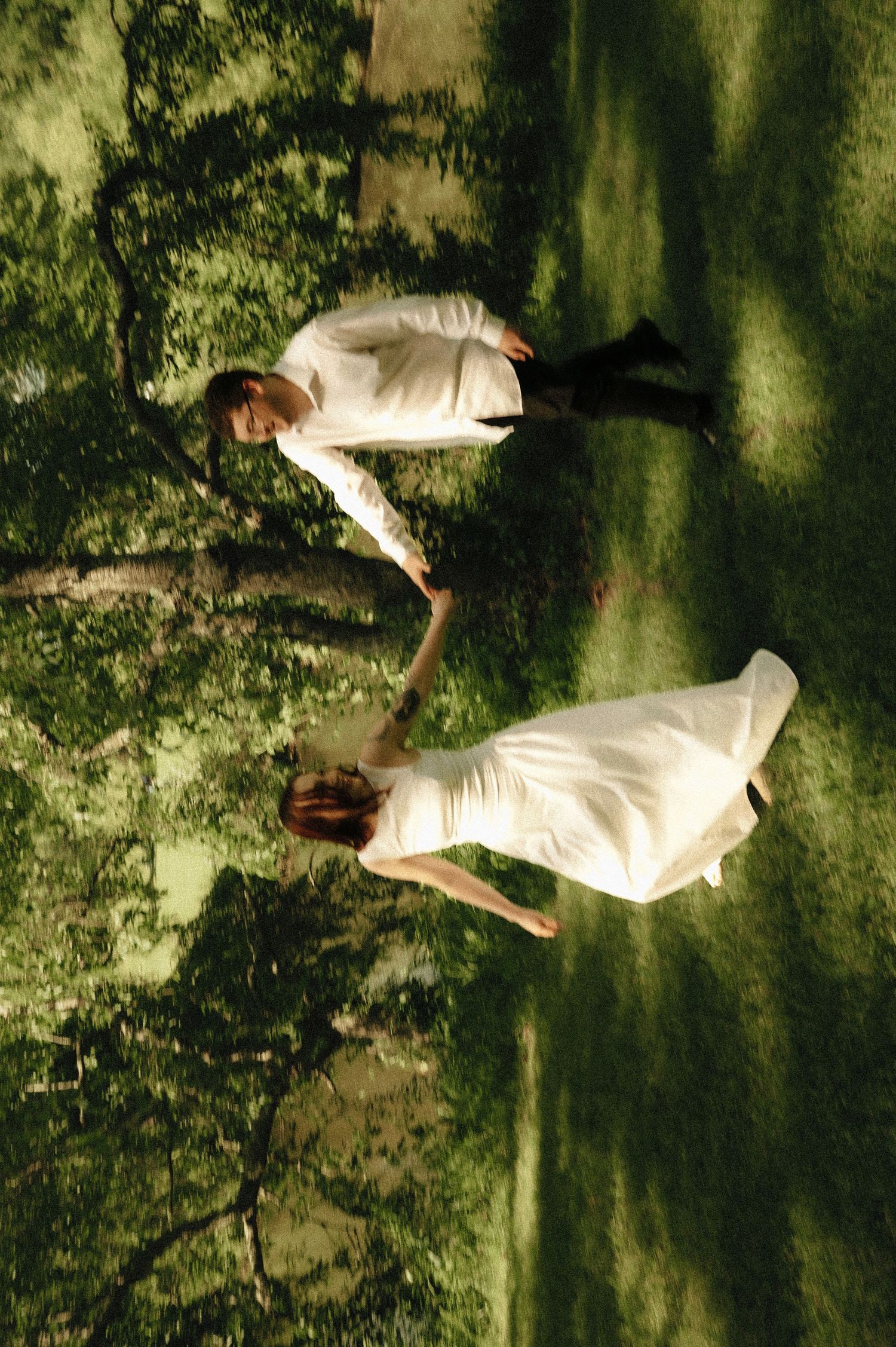 Aerial view of a couple in white wedding attire holding hands on a lush green lawn under tree shadows.