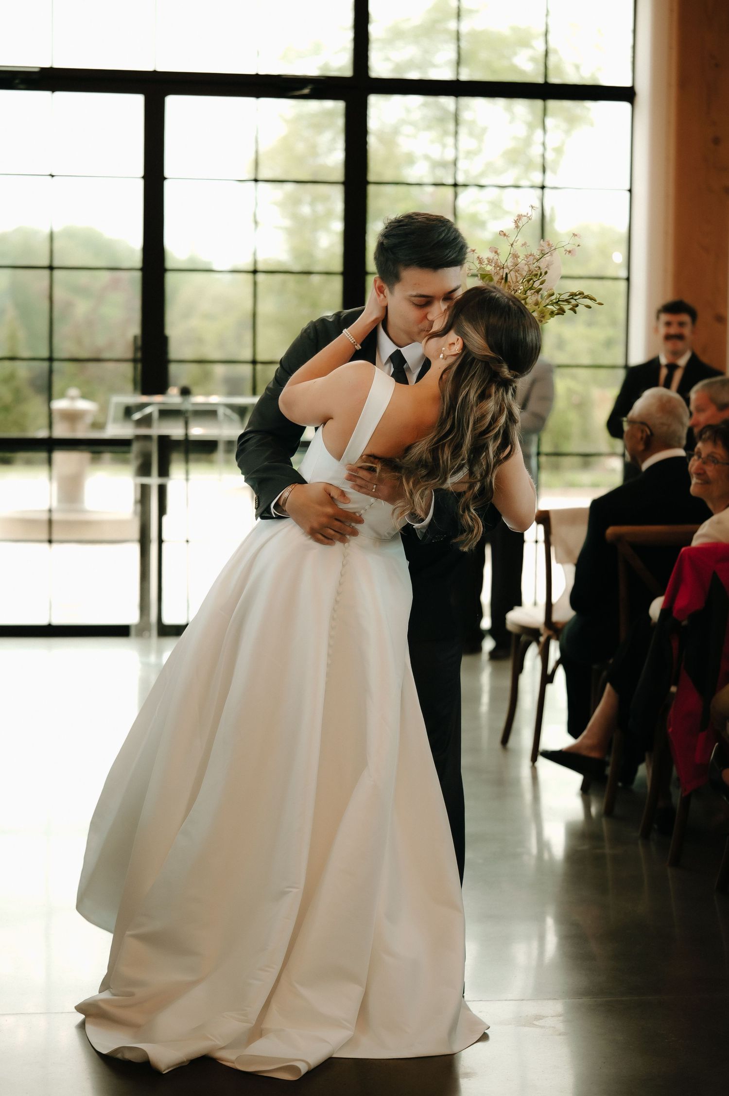 Bride and groom share first kiss in elegant indoor wedding venue with large windows.