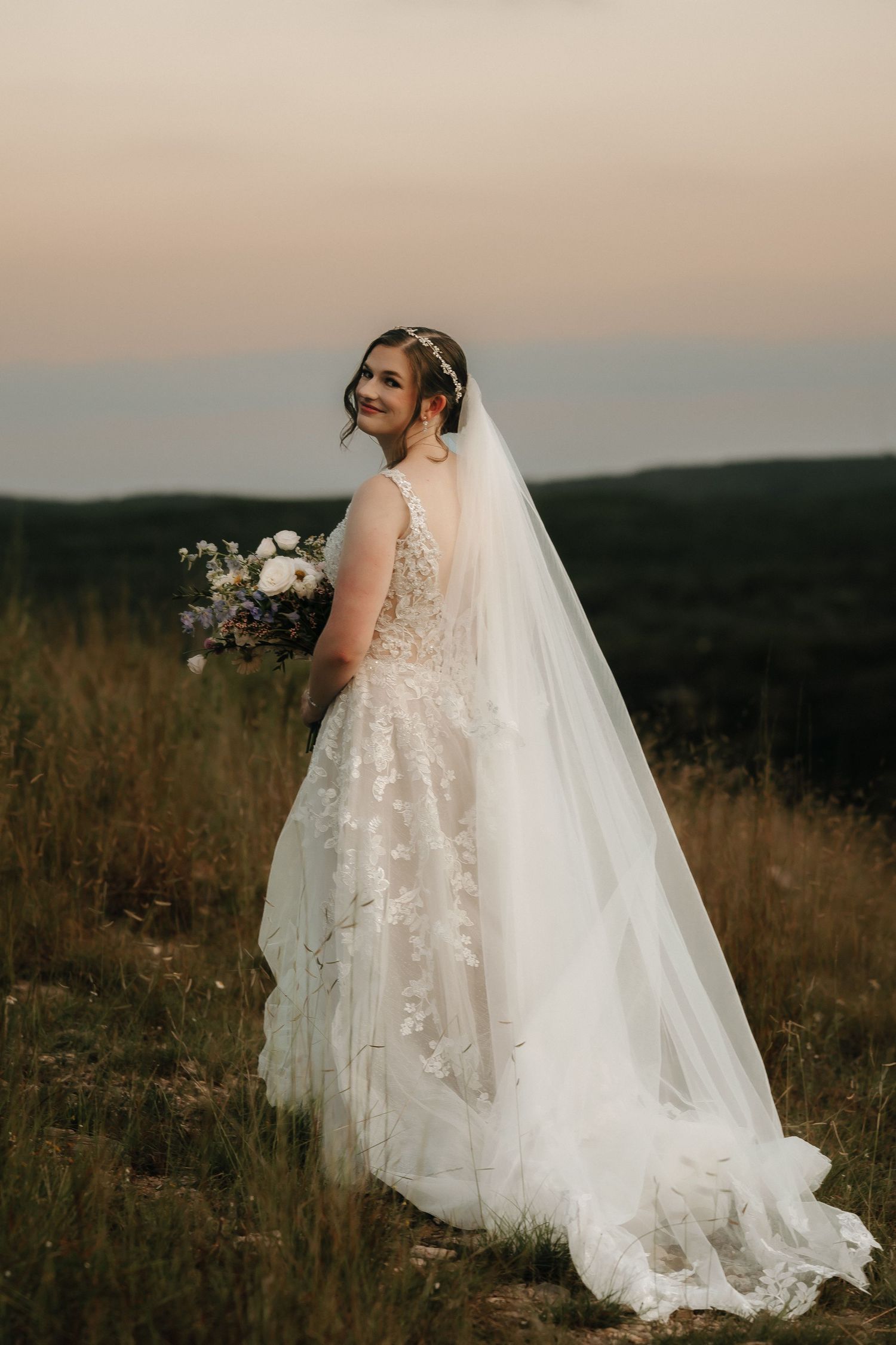 Bride in lace wedding gown with flowing train poses against sunset landscape.