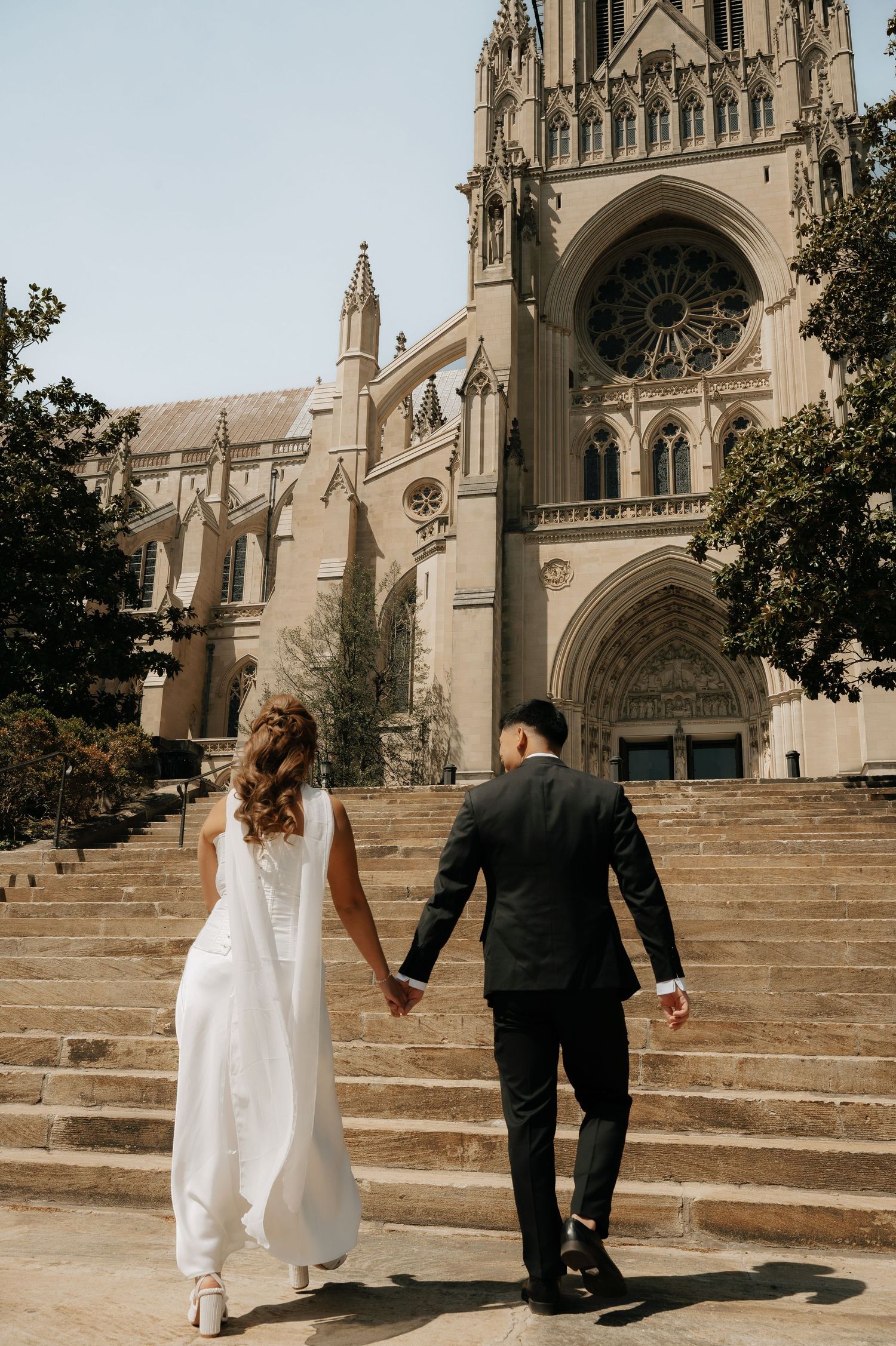 Bride and groom holding hands walking up stone steps toward a grand Gothic cathedral on their wedding day.